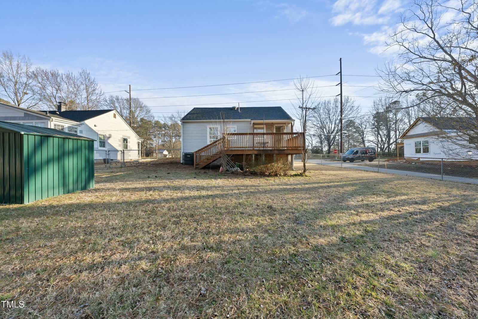 A small house with a raised wooden deck and steps, a large grassy backyard, a green shed on the left, and neighboring houses and trees in the background under a blue sky.