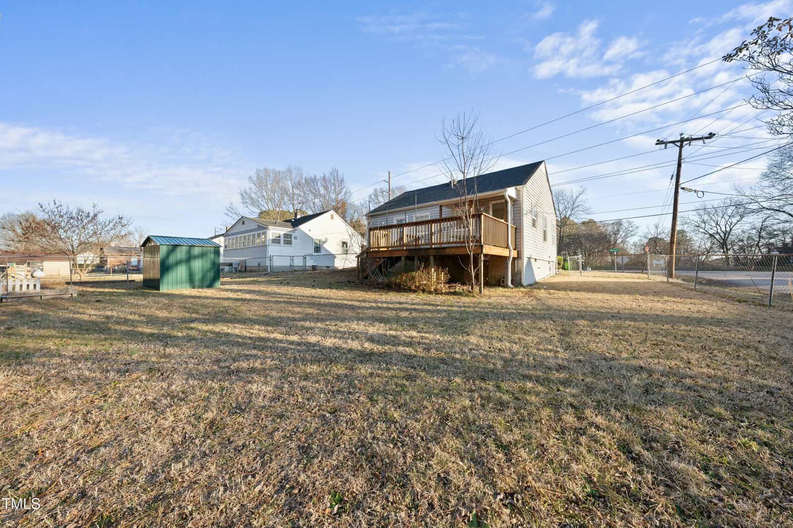 A backyard with dry grass, a small green shed, and a medium-sized house featuring a raised wooden deck. Power lines run along the side, and neighboring homes are visible in the background under a clear sky.