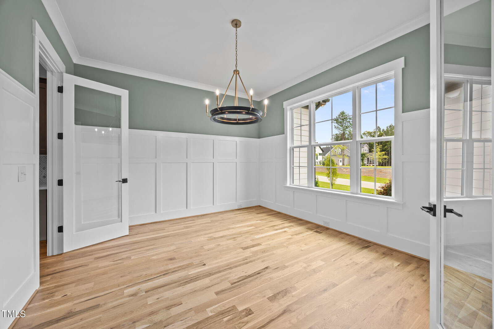 A bright, empty room with light wood floors, white wainscoting, pale green upper walls, a modern ring chandelier, large window showing a lawn and street, and double glass doors.