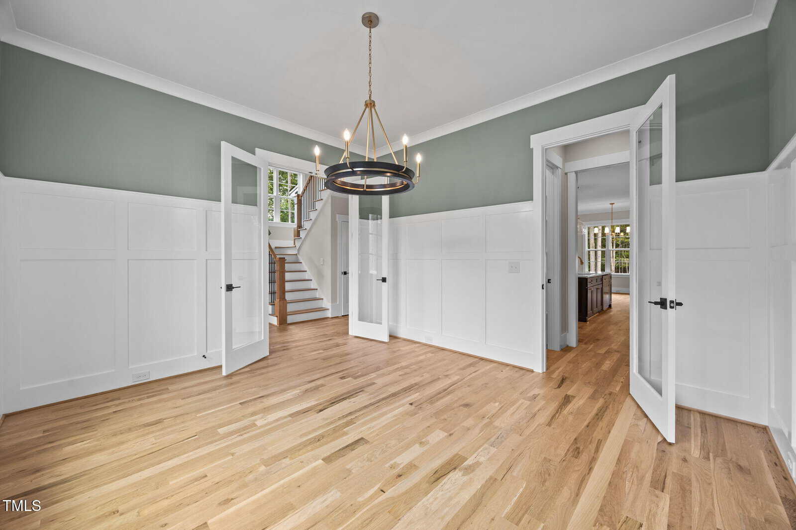A room with light wood floors, sage green and white paneled walls, a modern chandelier, double glass doors, visible staircase, and an adjoining room with large windows and a kitchen island.