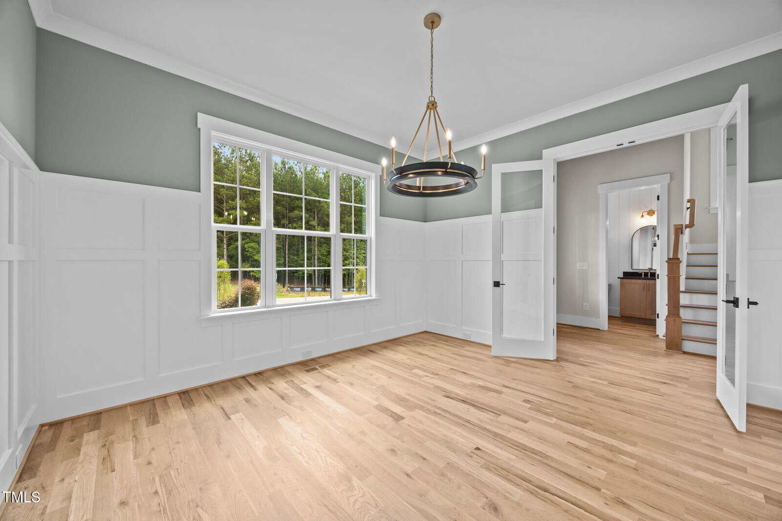 Empty room with light wood floors, white paneled walls, green upper wall section, large window, modern chandelier, double glass doors, and a view into a hallway with stairs and a bathroom.
