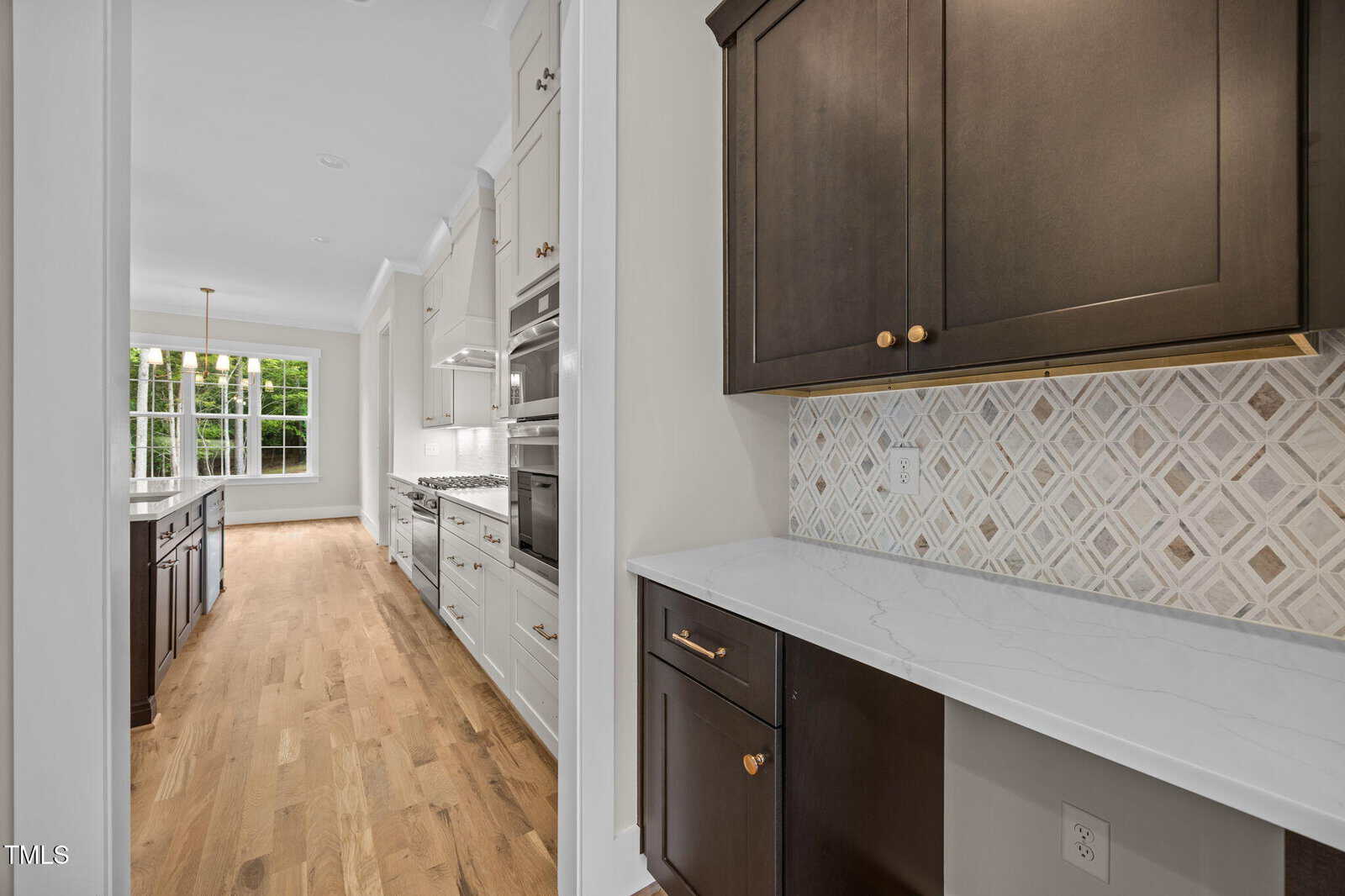 A modern kitchen with white cabinets, a gas stove, wood flooring, and a large window at the far end. In the foreground, a counter area features dark cabinets and a geometric tile backsplash.