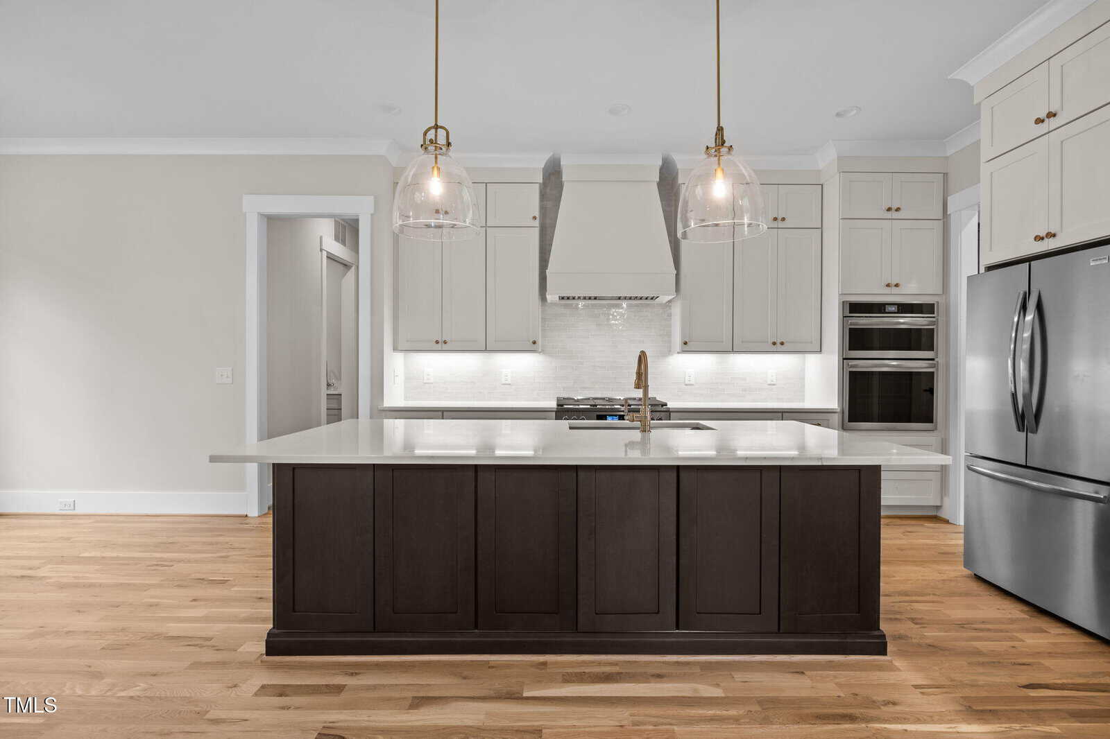 Modern kitchen with white upper cabinets, stainless steel appliances, a dark island with a light countertop, two pendant lights, wood flooring, and a white backsplash. The space is well-lit and appears clean and new.