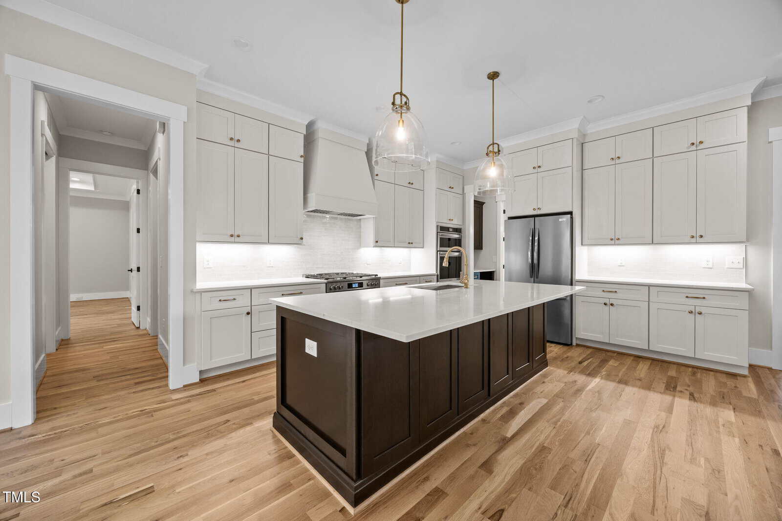 A modern kitchen with light cabinets, a dark island, stainless steel appliances, two pendant lights, a built-in stove, wood floors, and white countertops. The room is well-lit and has a clean, minimalist design.