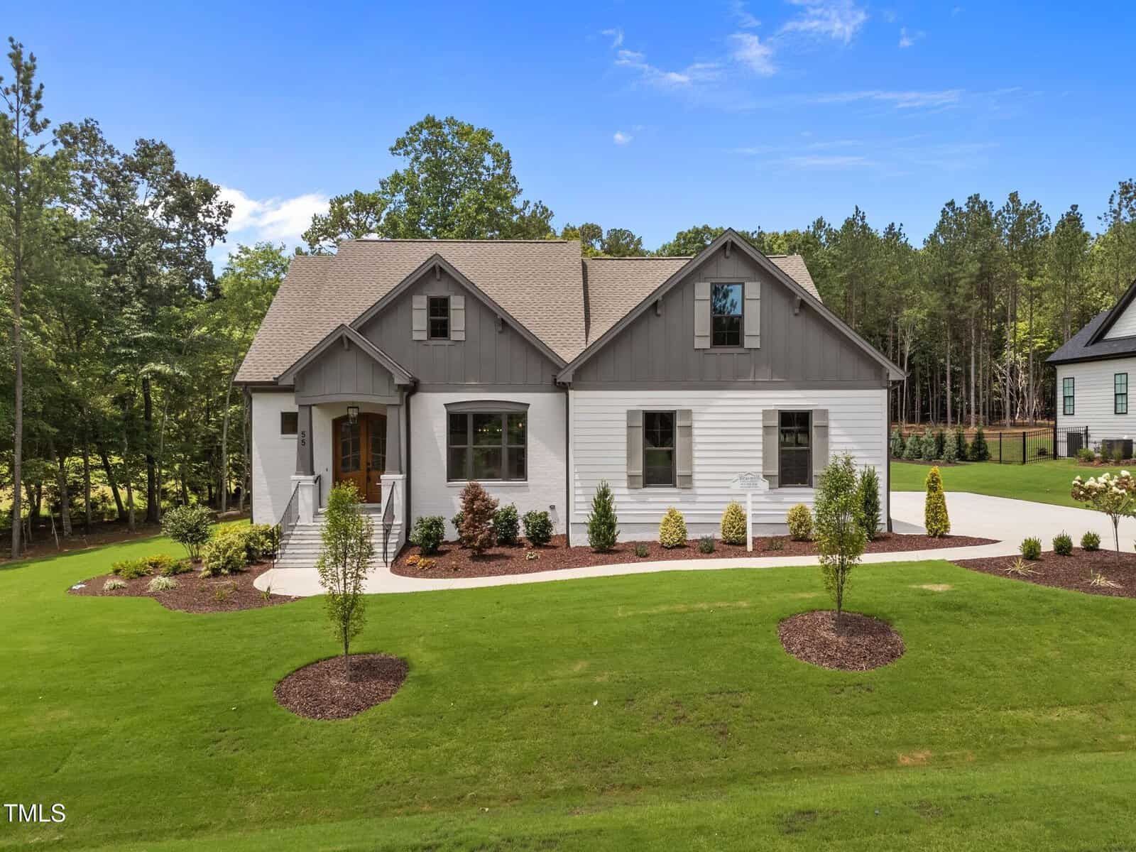 A modern farmhouse-style home at 55 Cherry Bark Drive in Youngsville features a gray gabled roof, white and gray siding, and a landscaped front yard. The house sits on a large, manicured lawn bordered by woods under a partly cloudy blue sky.