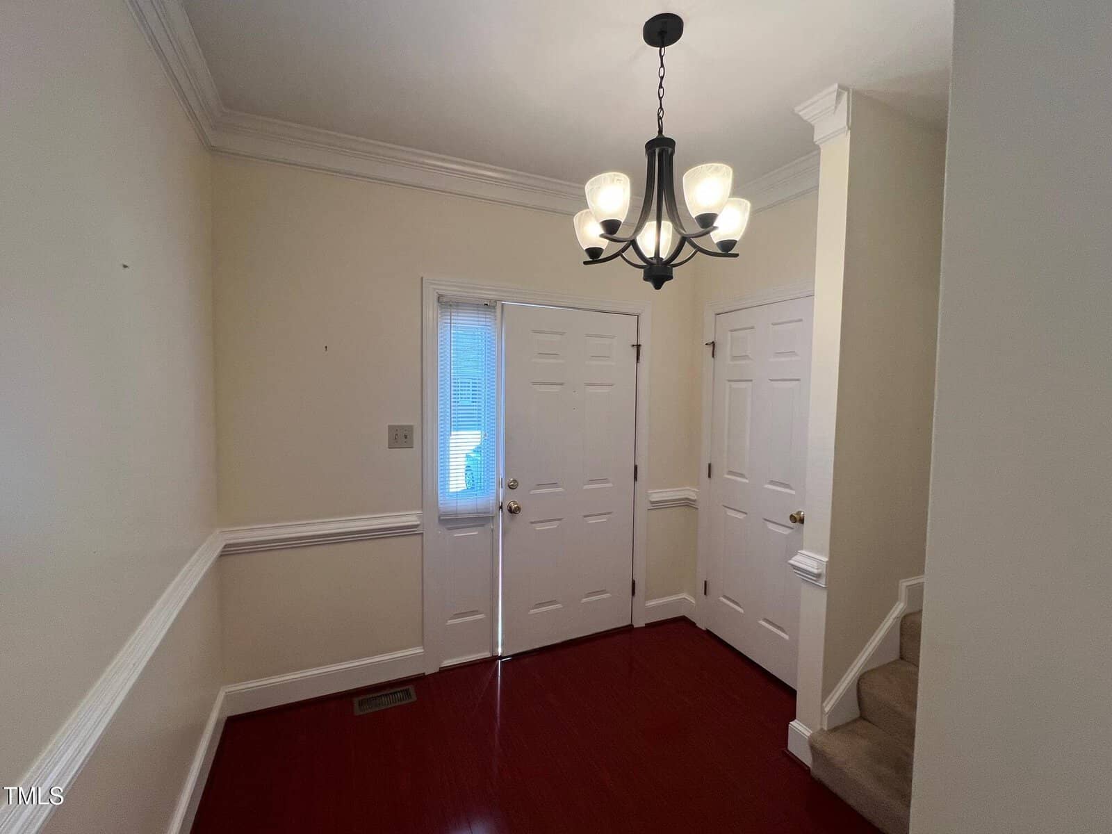 A small, empty foyer with hardwood floors, cream-colored walls, and white trim. There are three white doors, a small staircase with beige carpet, and a black chandelier with frosted glass shades hanging from the ceiling.