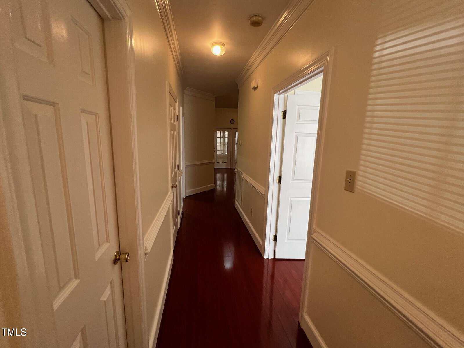 A hallway with polished wooden floors, cream-colored walls, and white doors. The ceiling has a light fixture, and molding decorates the top and middle of the walls. One door is partially open, leading to another room with a window.