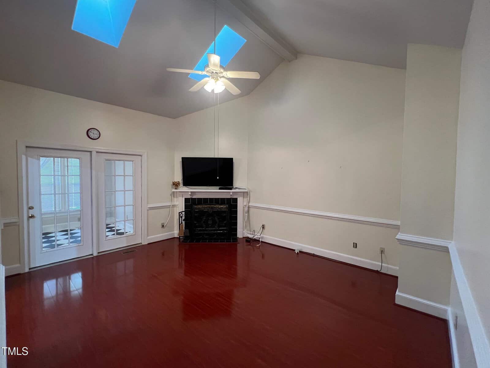 A living room with high ceilings featuring a ceiling fan and two skylights. A fireplace with a black surround is centered on the far wall, with a flat-screen TV above it. Double doors with glass panes lead to another room with a checkered floor.