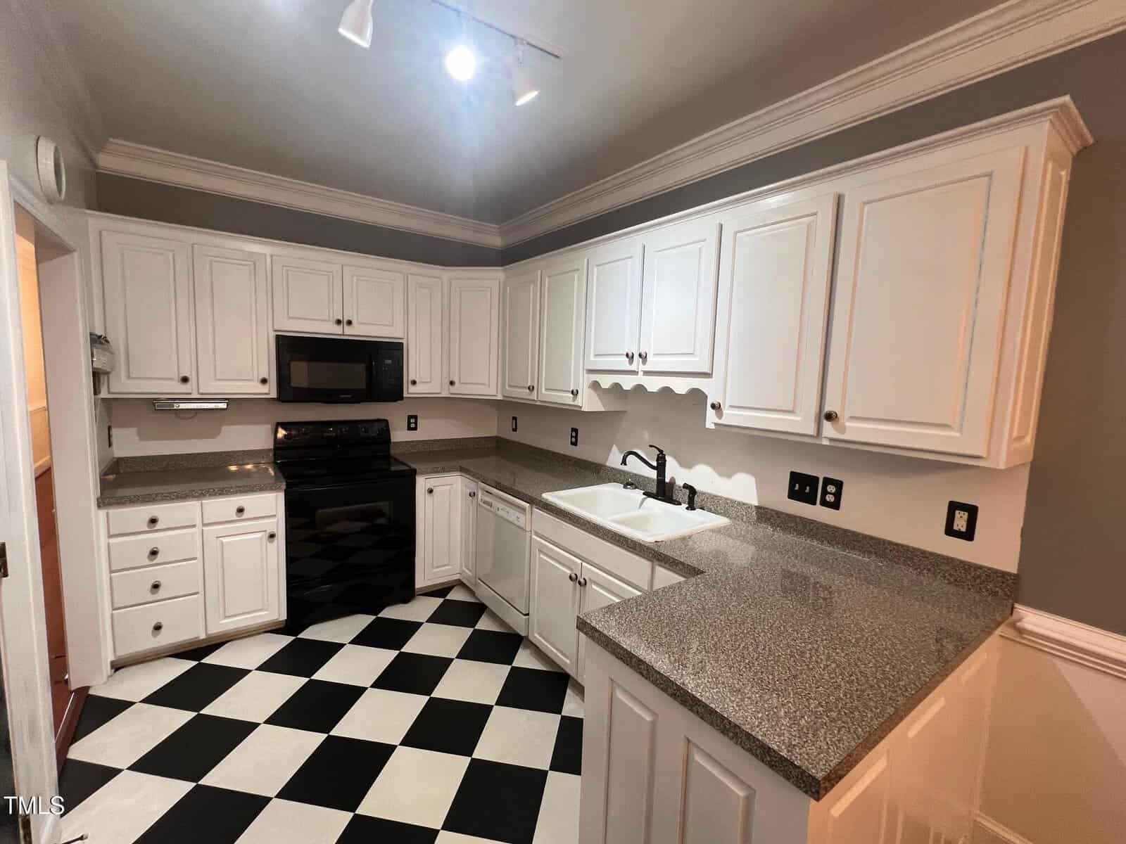 A kitchen with white cabinets and dark gray countertops. It features a black oven, stove, and microwave. The flooring is black and white checkerboard. The walls are a muted gray, and there's track lighting on the ceiling.