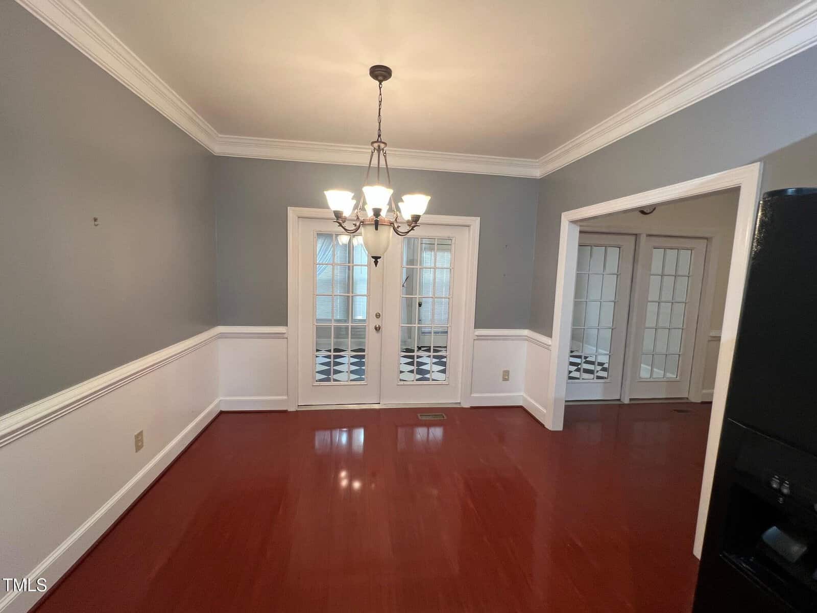 A dining room with wooden flooring and gray walls, featuring a chandelier hanging from the ceiling. Double French doors open to another room with black and white checkered flooring. A portion of a black refrigerator is visible on the right.