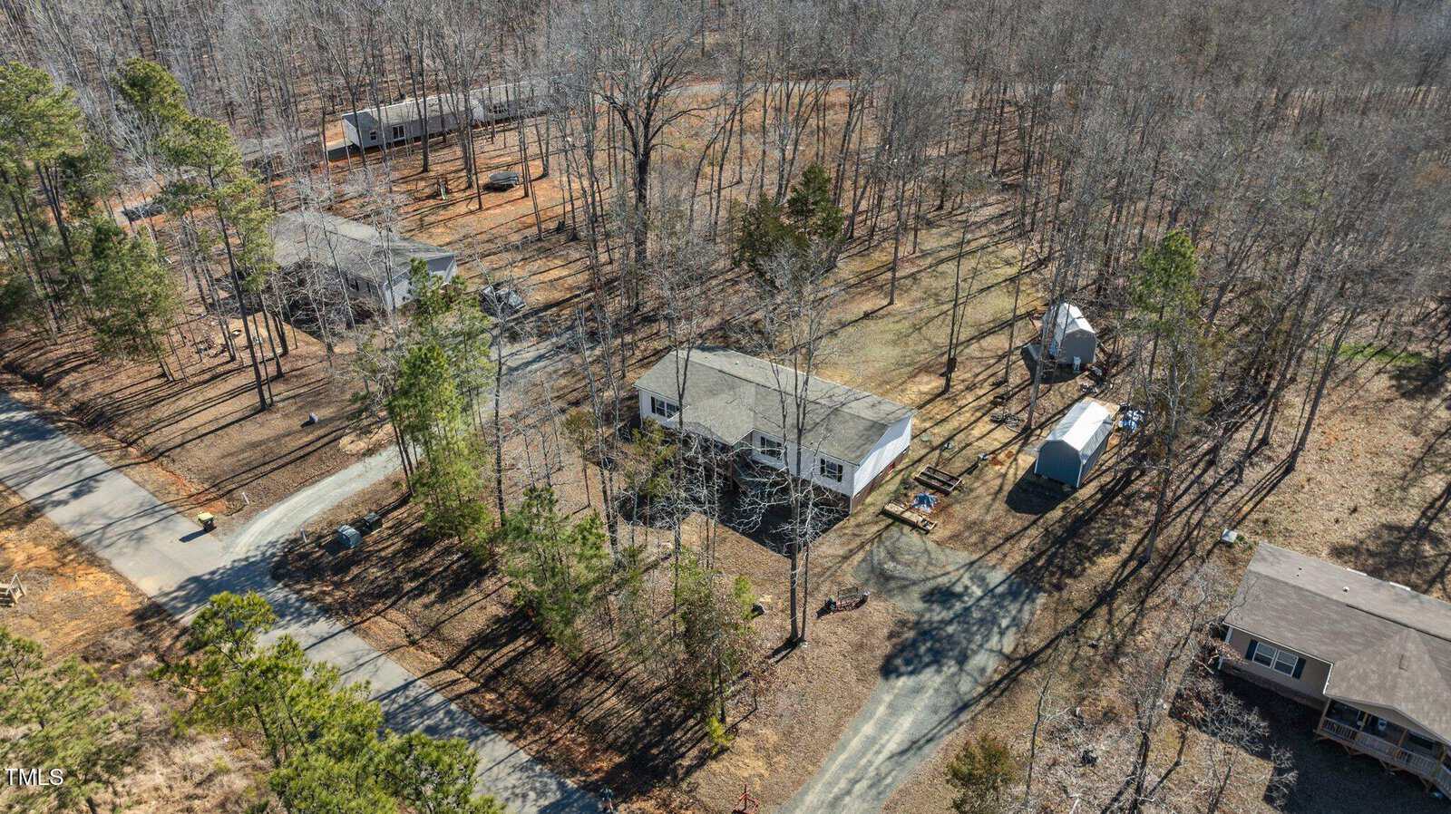 Aerial view of Oxford's scenic residential area at 6636 Woodlawn Drive, surrounded by leafless trees. Several houses dot the landscape with a large metal structure nearby. The gravel road winds through wooded areas, where shadows from the bare trees softly blanket the ground.