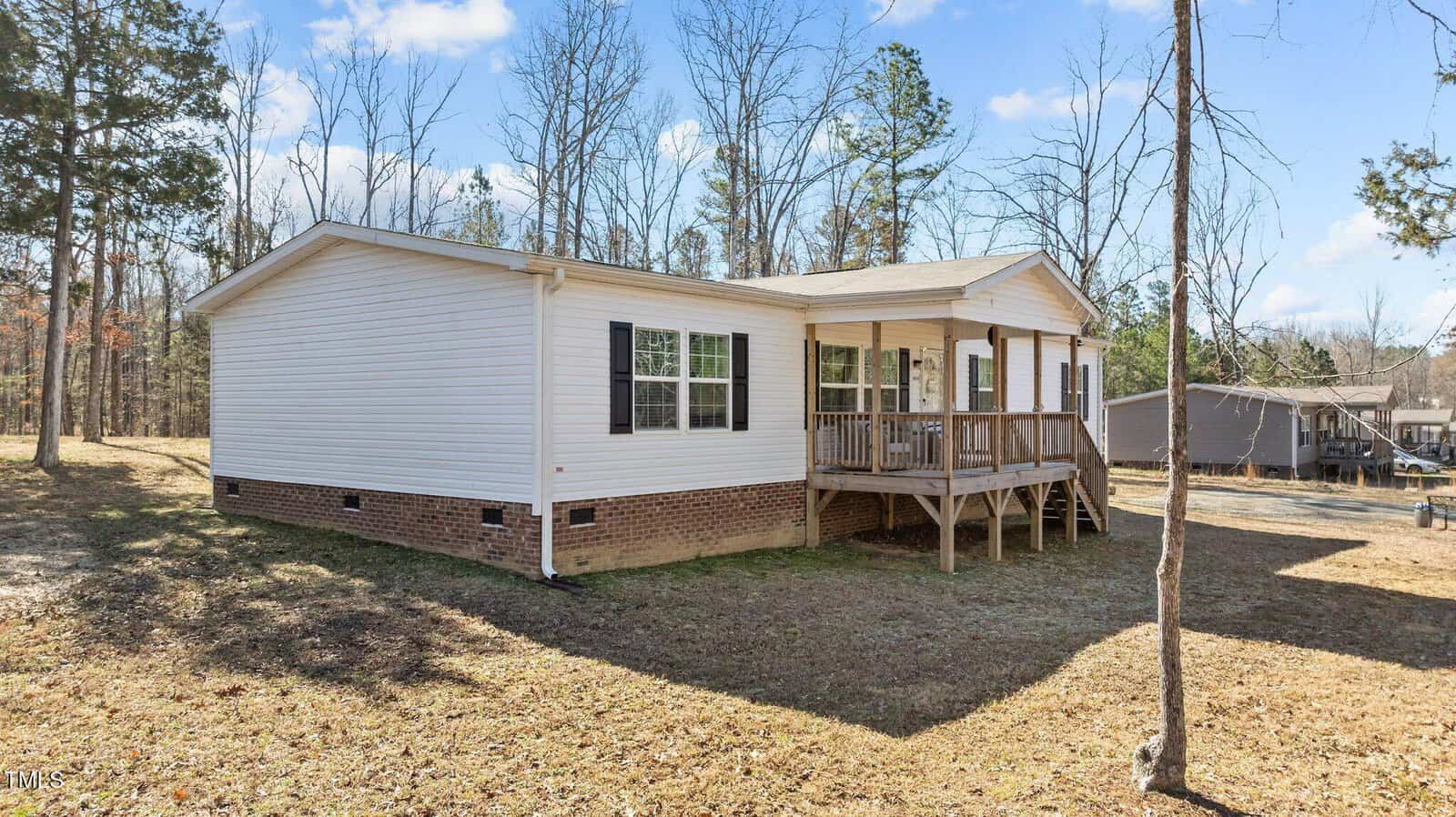 A single-story manufactured home with light-colored siding and a brick foundation sits on a grassy lot with sparse trees. The home features a small front porch with a railing and steps leading to the front door. The sky is clear and sunny.
