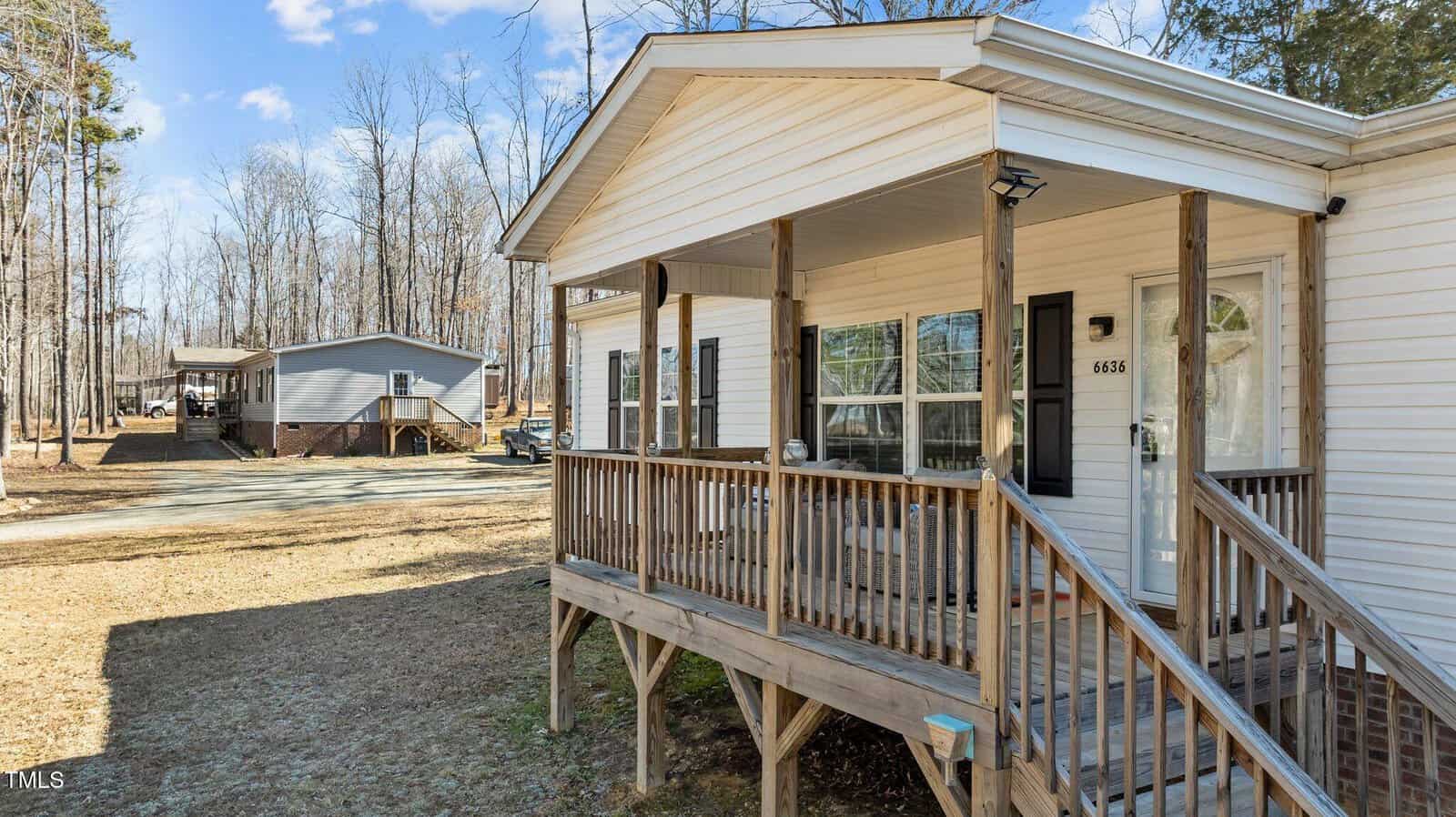 A white, single-story house with a wooden porch and steps leading up. The porch has railings and a light near the door. Another similar house is visible in the background amid leafless trees under a clear blue sky.