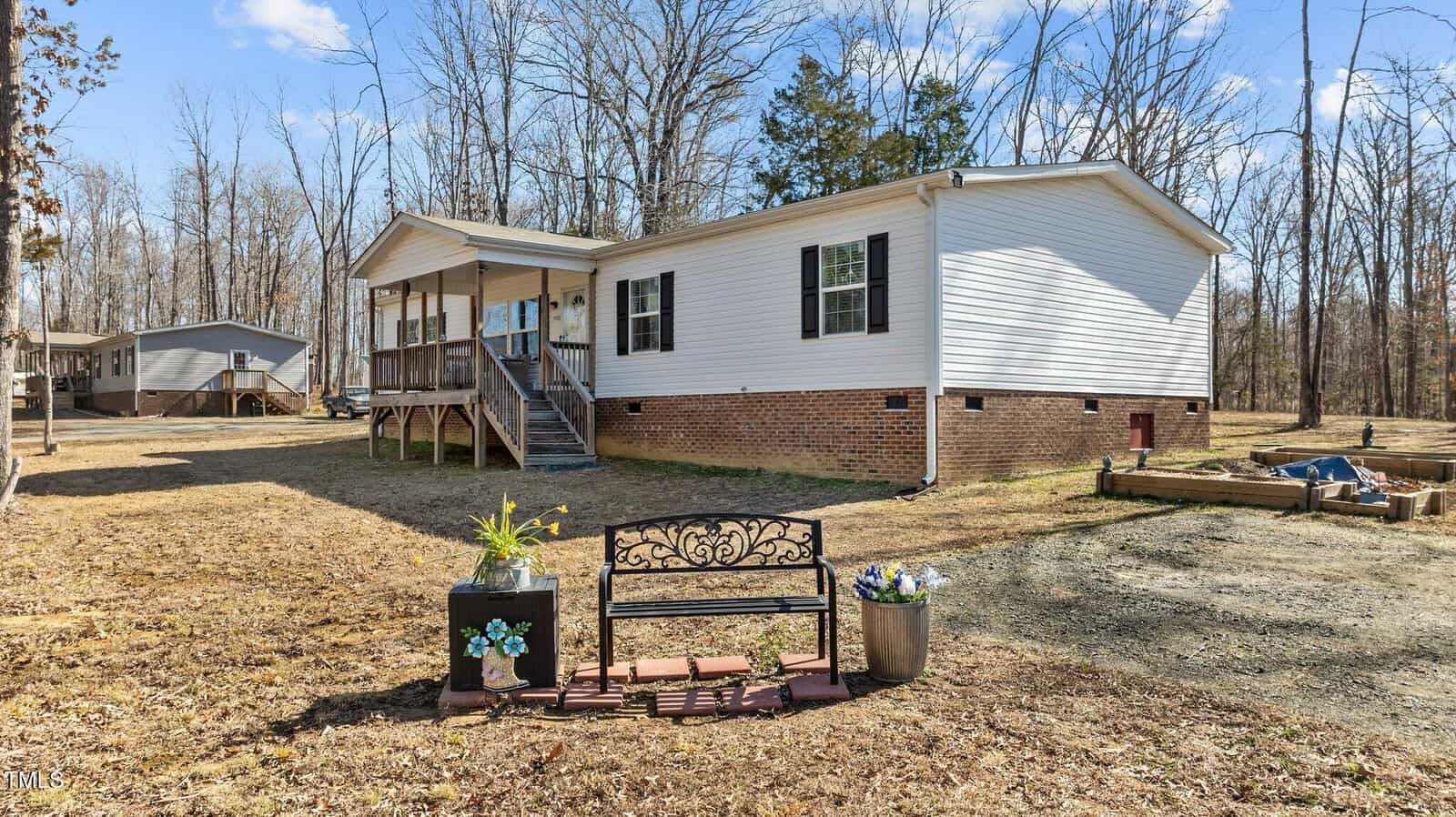A white manufactured home with a brick foundation is set in a wooded area. A front porch with steps leads to the entrance. In the foreground, there's a black metal bench, a potted plant, and a small raised garden bed. Sparse trees surround the area.