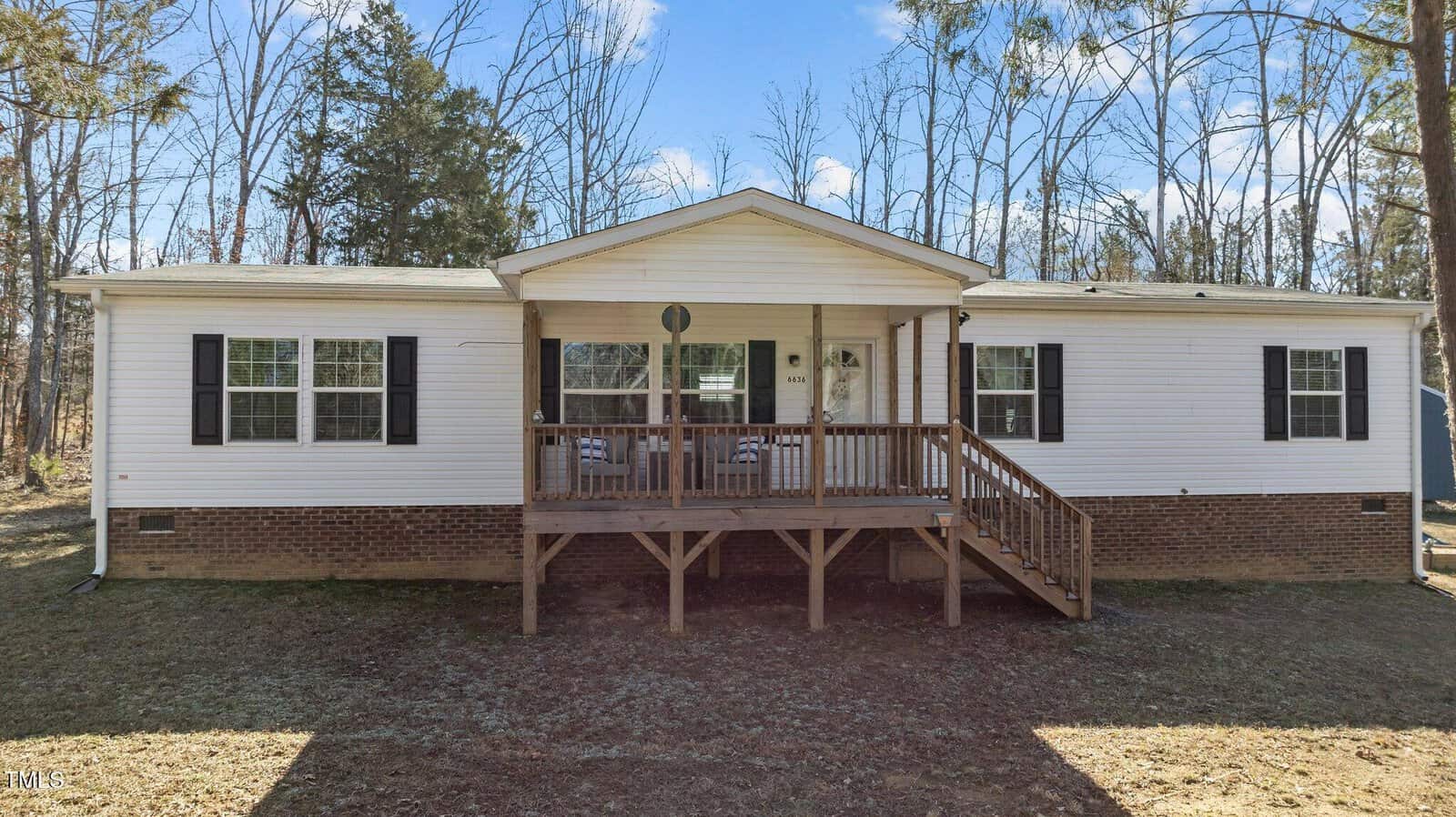 A single-story white house with black shutters and a brick foundation. It has a wooden porch with steps leading up to the front door. The house is surrounded by trees, and the sky is clear and sunny.