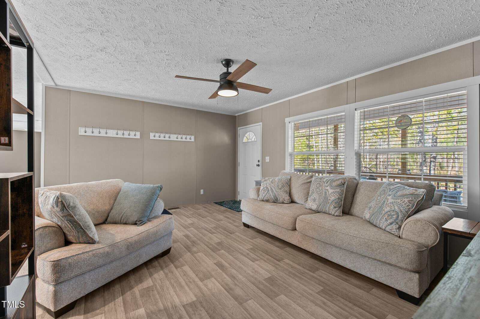The image shows a living room with two beige sofas facing each other. There are decorative pillows on each sofa. A ceiling fan with a light fixture is centered overhead. Large windows with blinds are on the right wall, and the floor is wood-patterned.