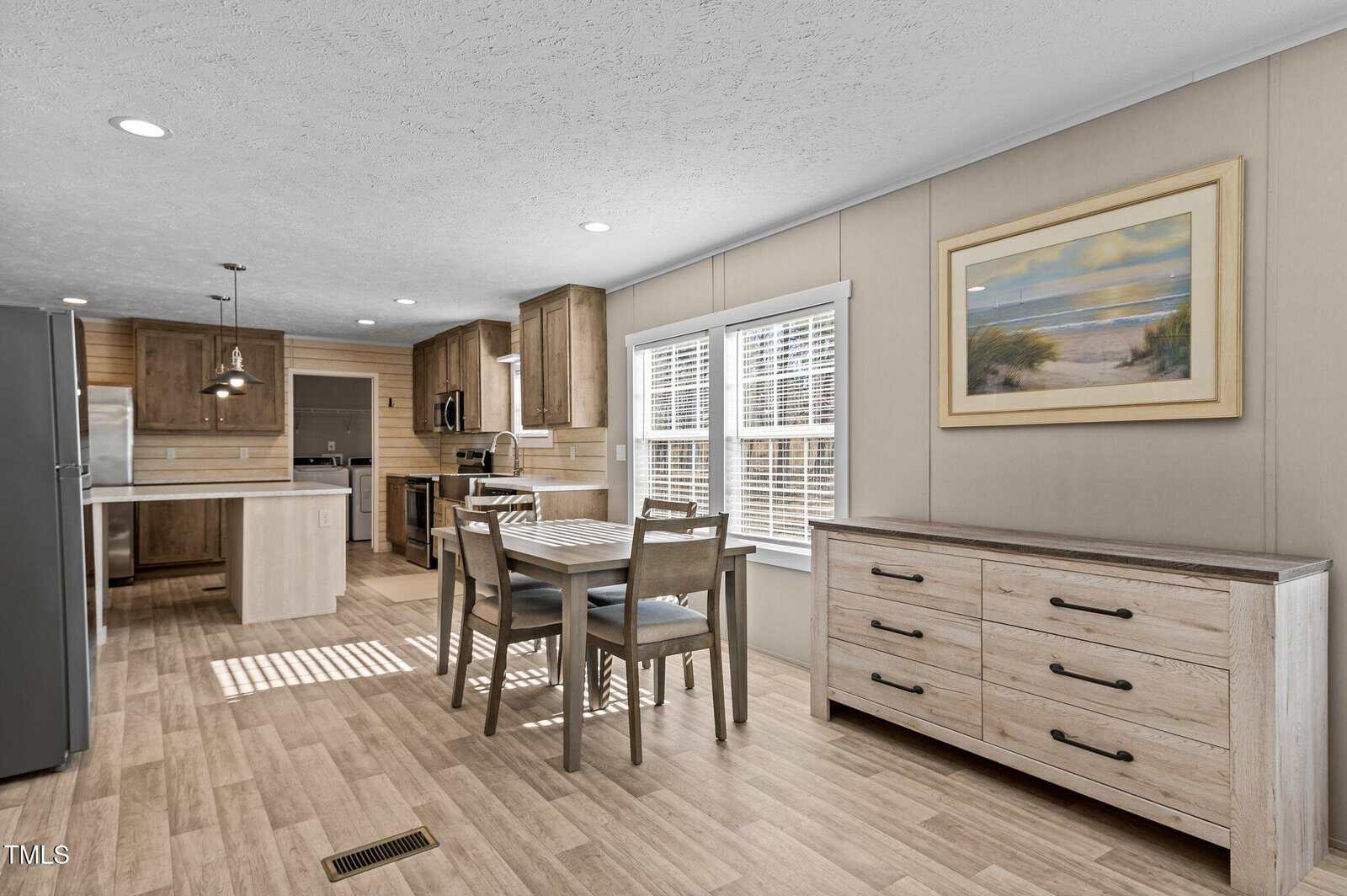 A modern kitchen and dining area with light wood flooring and cabinetry. The room features a dining table with four chairs, a large island, stainless steel appliances, and a landscape painting on the wall. Sunlight filters through the window blinds.