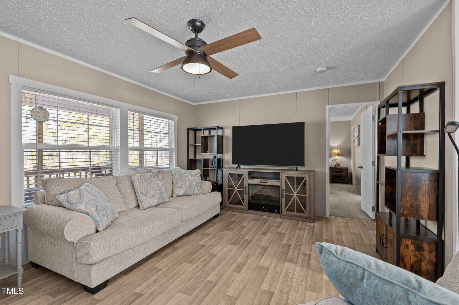 A living room featuring a beige sofa with patterned cushions on a wooden floor. A large flat-screen TV is mounted on a stand with cabinets. The room has a ceiling fan, two tall shelves, and large windows with blinds on the left. A doorway leads to another room.