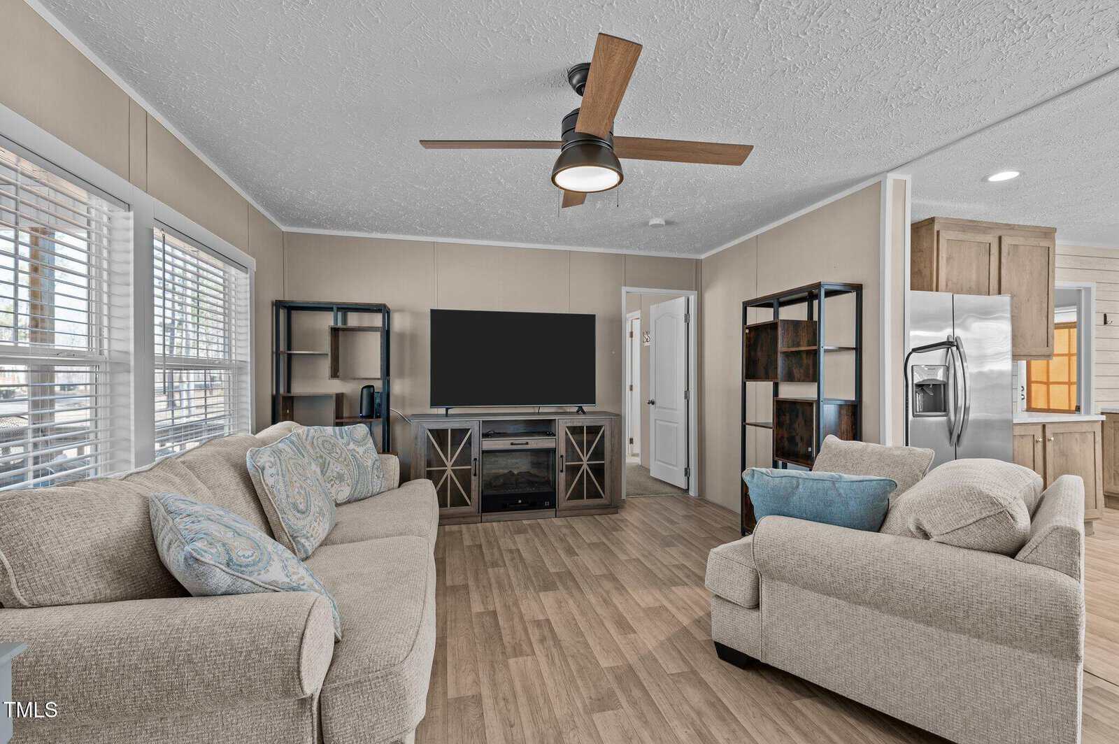 A living room with a beige sofa and loveseat, a large flat-screen TV on a console, two shelving units, and a ceiling fan. The room has wood-look flooring, beige walls, and multiple windows with blinds. A stainless steel refrigerator is visible in the background.