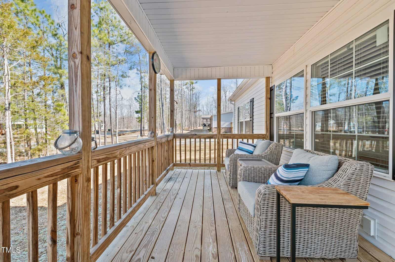 Front porch with wooden flooring and railing, featuring two wicker chairs with cushions and a small wooden side table. The porch is covered with a white ceiling, and the background shows trees and a nearby building.