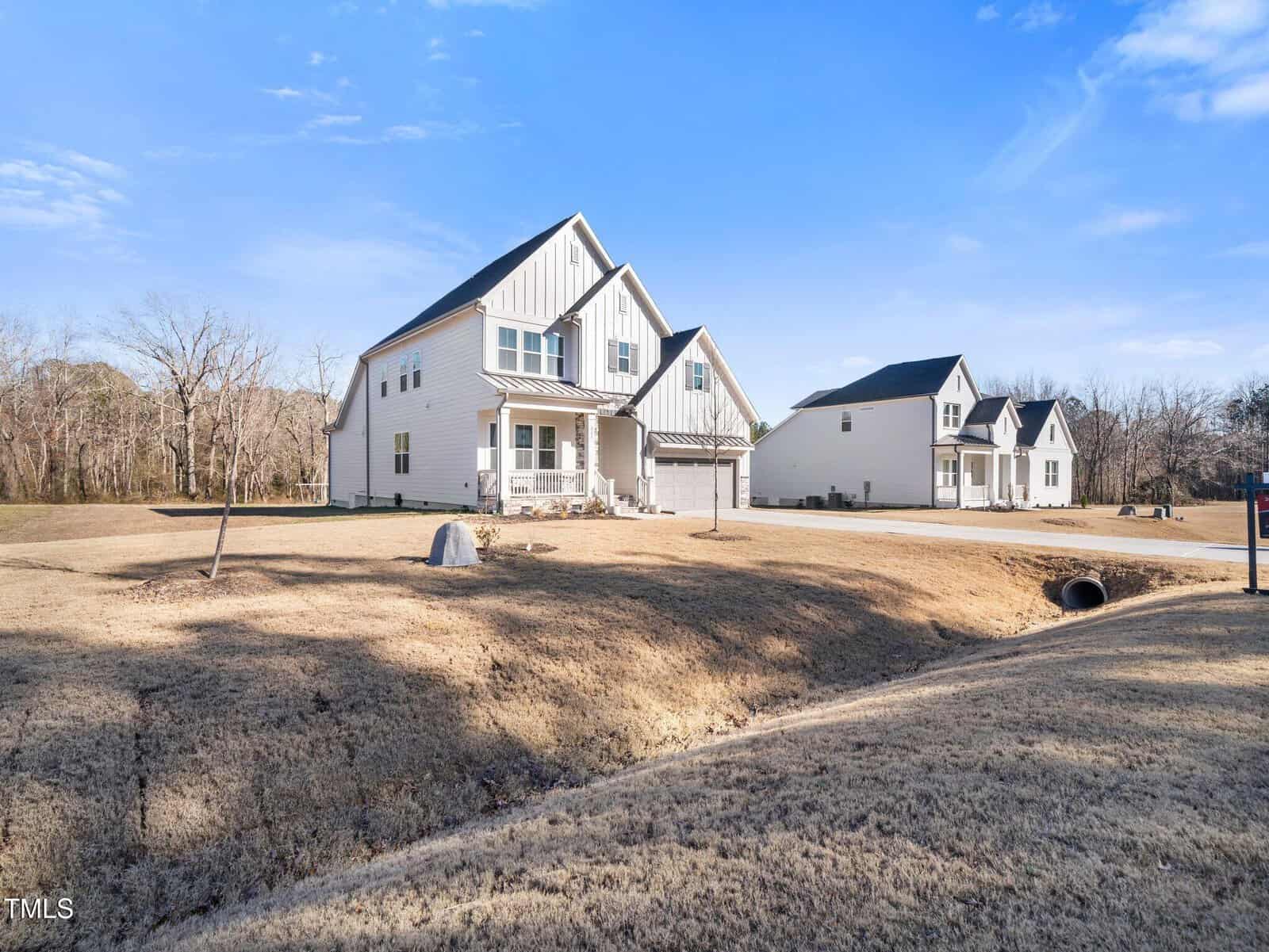 A modern white two-story house with a covered front porch, attached garage, and large yard, set in a suburban neighborhood with a similar house visible in the background under a clear blue sky.