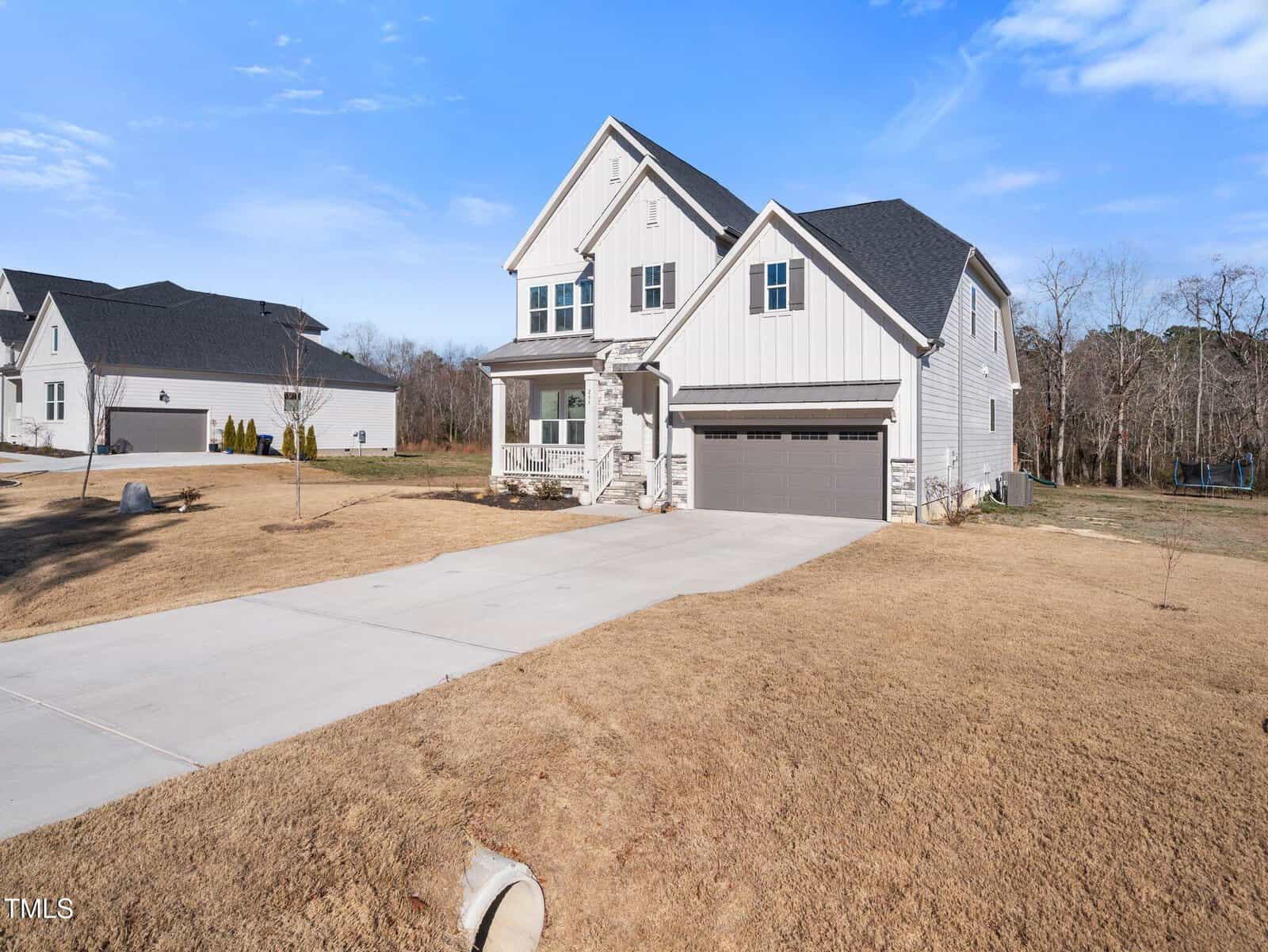 A modern two-story house with white siding, a gray roof, and a two-car garage sits on a large lot with dry, brown grass. A long concrete driveway leads to the garage. Blue sky and trees are in the background.