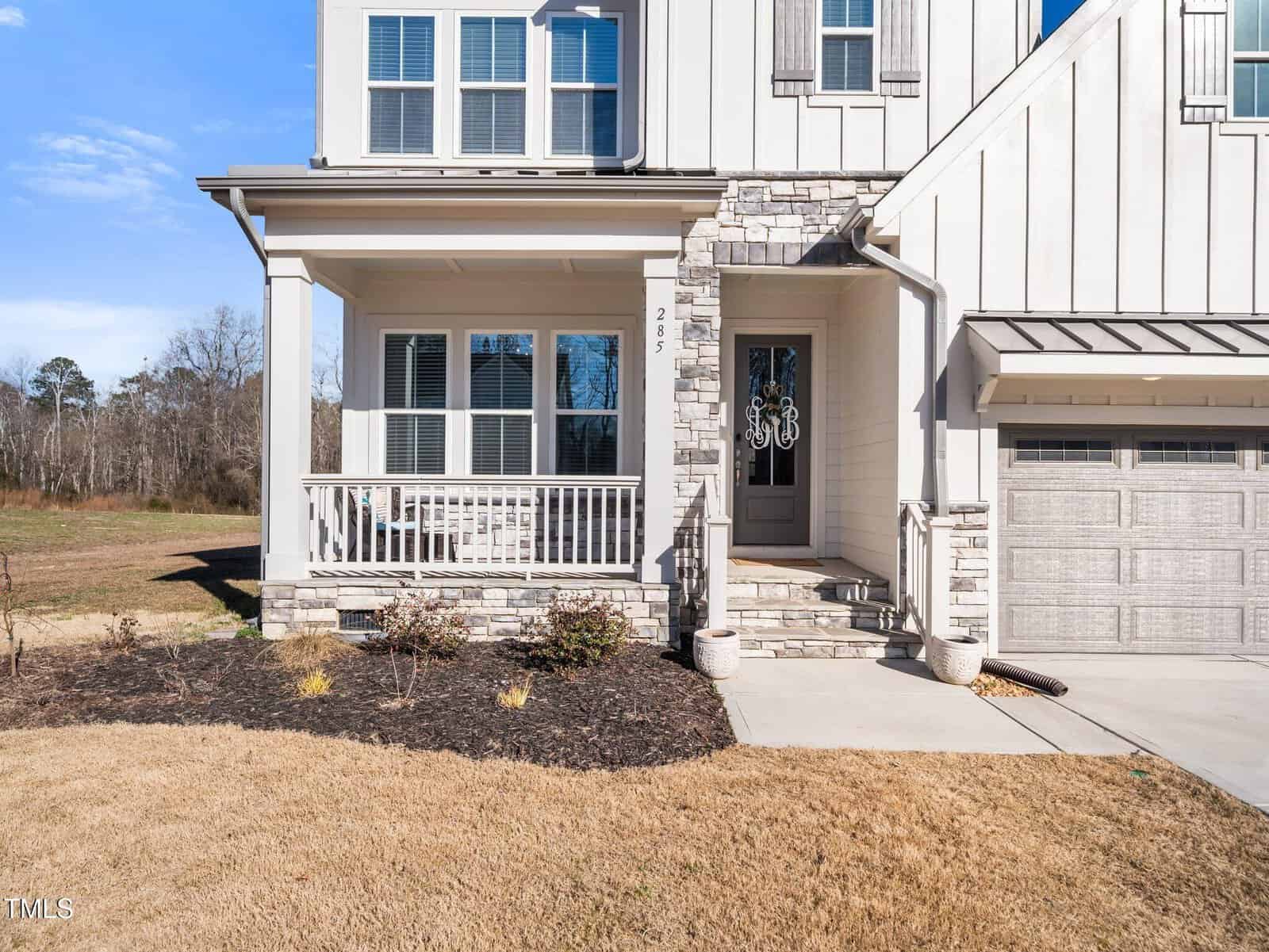 A two-story house with white siding and stone accents, featuring a covered front porch with a bench, steps leading to a decorative front door, and a two-car garage on the right. The yard has dry grass and sparse landscaping.
