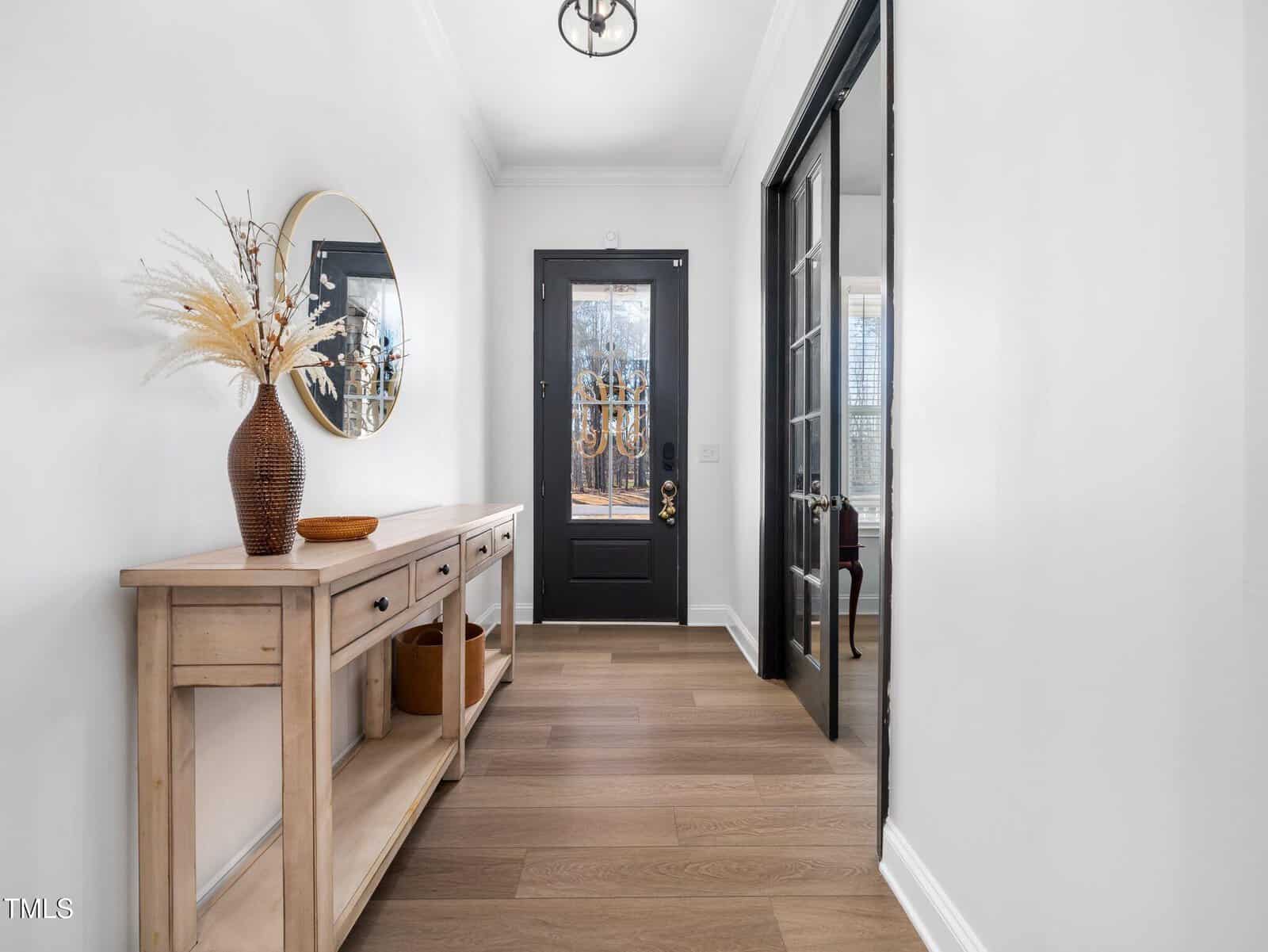 A hallway with light wood flooring, a wooden console table with a brown vase holding dried plants, a round mirror on the left wall, and a black front door with glass panels at the end of the hall.
