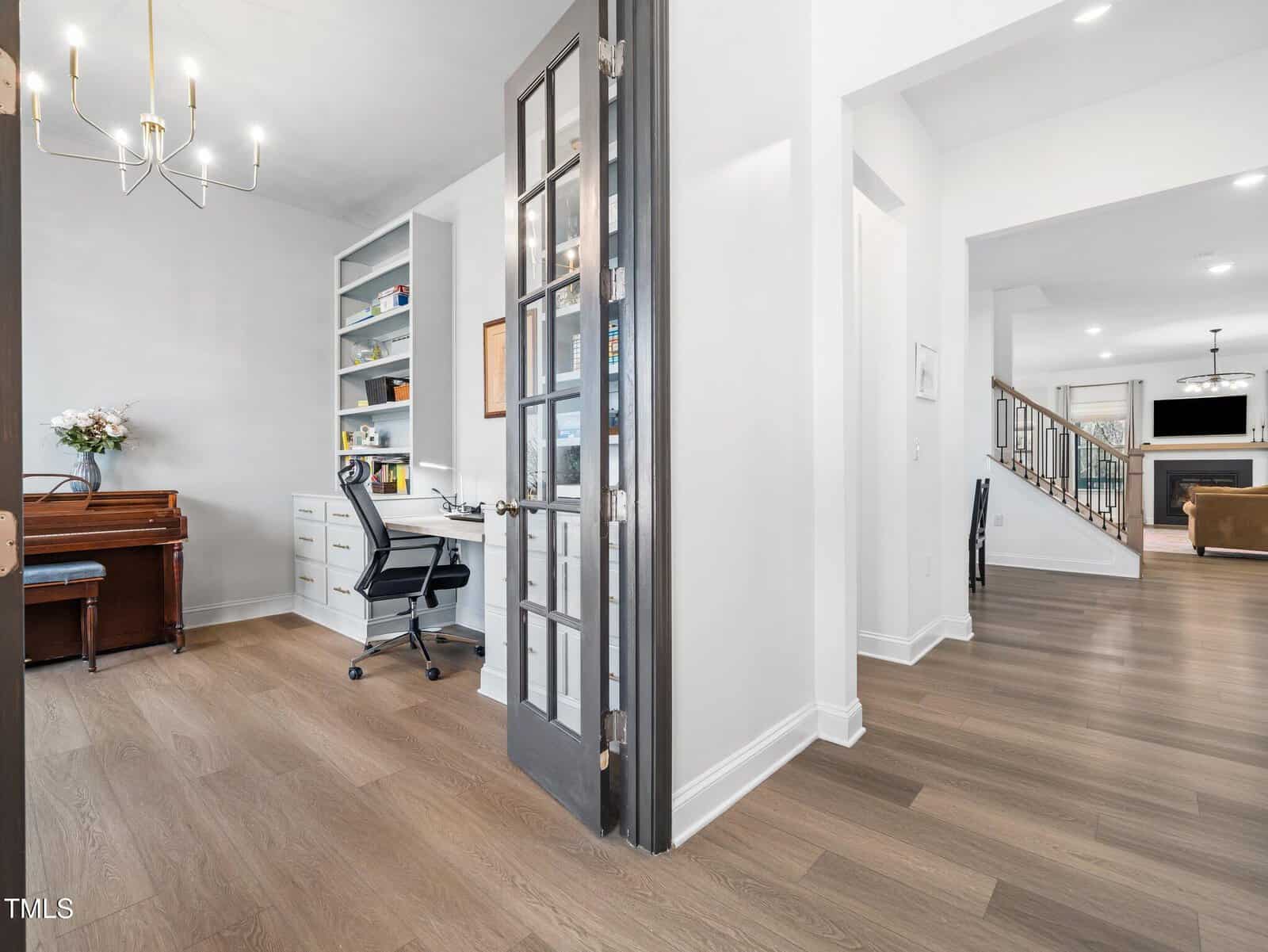 A modern home office with a desk, shelves, and chair is separated by glass French doors from a hallway leading to a living area with a fireplace, staircase, and brown sofa. A piano is against the office wall.