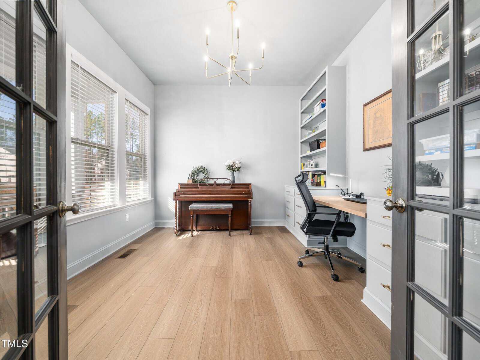 A bright home office with wood floors, a piano against the back wall, a desk with an office chair, built-in shelves, large windows on the left, and glass double doors in the foreground.