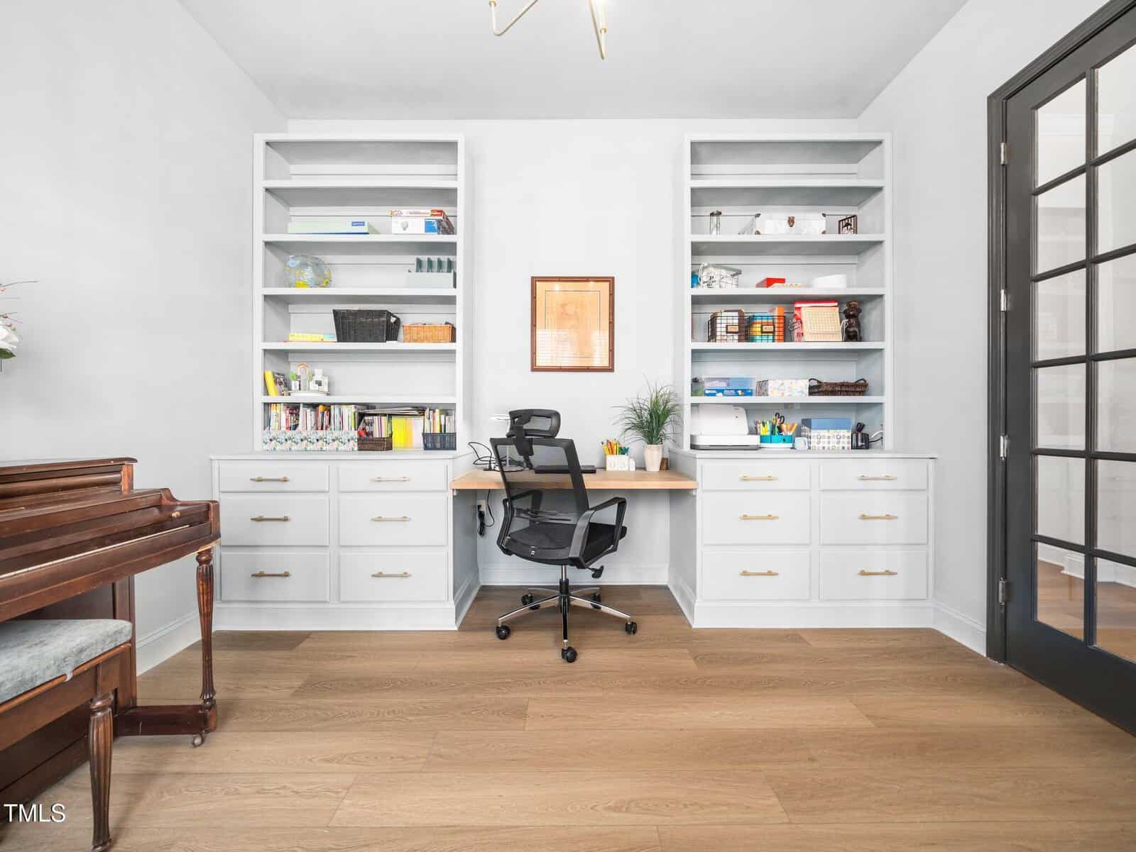 A home office with white built-in shelves, a wooden desk with a black office chair, books and plants on the shelves, a brown upright piano on the left, and light wood floors. Black-framed glass doors are on the right.