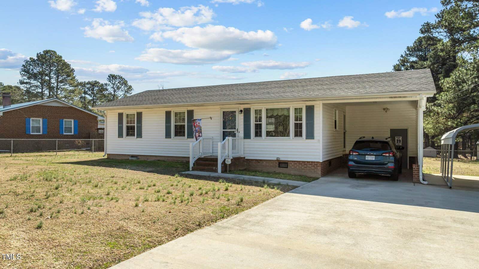A single-story white house on 1397 Ruin Creek Road boasts a brick foundation and a small porch. The driveway leads to a carport on the right with a parked car. The lawn is sparse, with some grass patches, and the neighboring Henderson home is partially visible on the left.