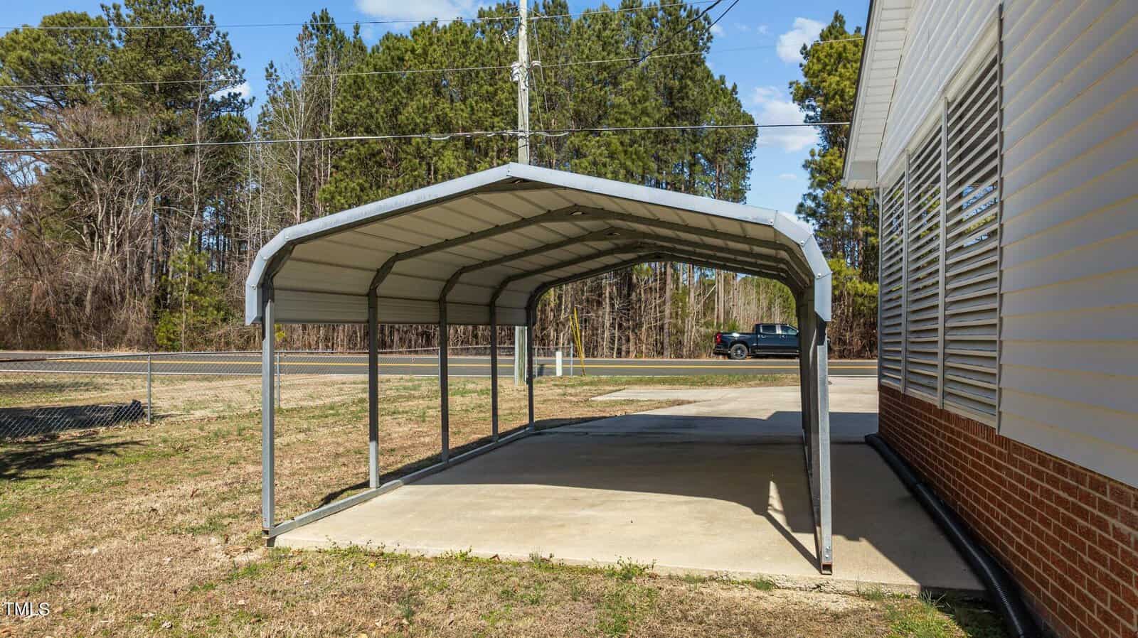 A metal carport with a curved roof stands next to a brick and siding building. It covers a concrete slab. In the background, a road is visible with a truck driving by, and there are trees lining the roadside.