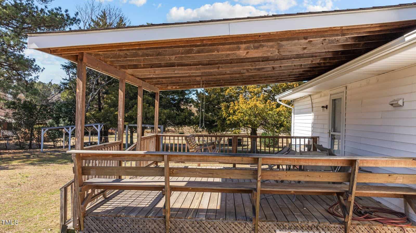 A wooden deck with a partial roof is attached to a white house. The deck has a railing and several patio chairs. The area is surrounded by trees and grass under a partly cloudy sky.