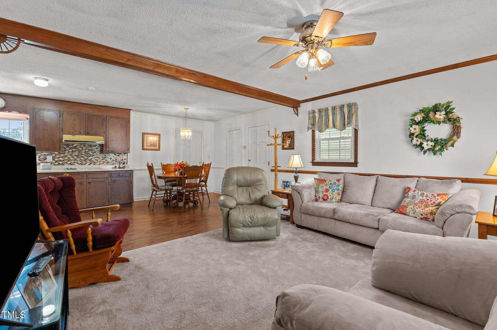 A cozy living room and dining area with a ceiling fan. The space includes a sofa, recliner, dining table with chairs, and a kitchen area with wooden cabinets. Decor includes a wall wreath, floral pillows, and a patterned curtain.