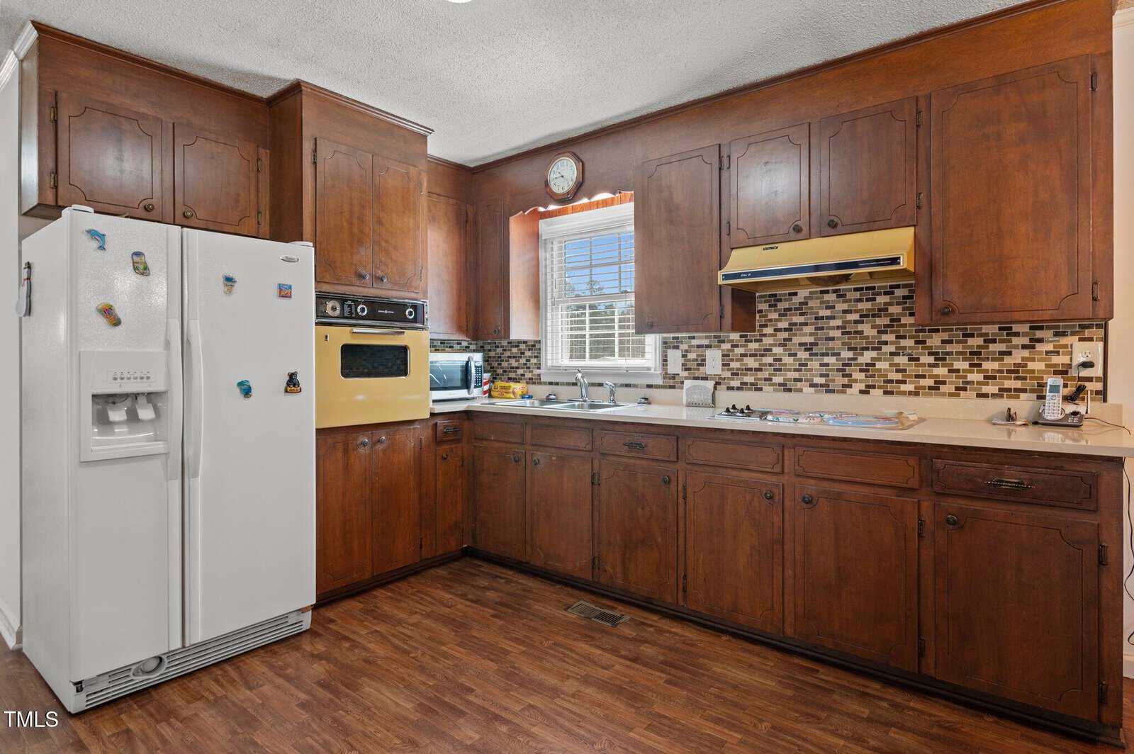 A vintage kitchen with dark wooden cabinets and a brown tile backsplash. Features include a white refrigerator with magnets, a yellow oven and range hood, a window above the sink, and a microwave on the counter. The floor is wooden.