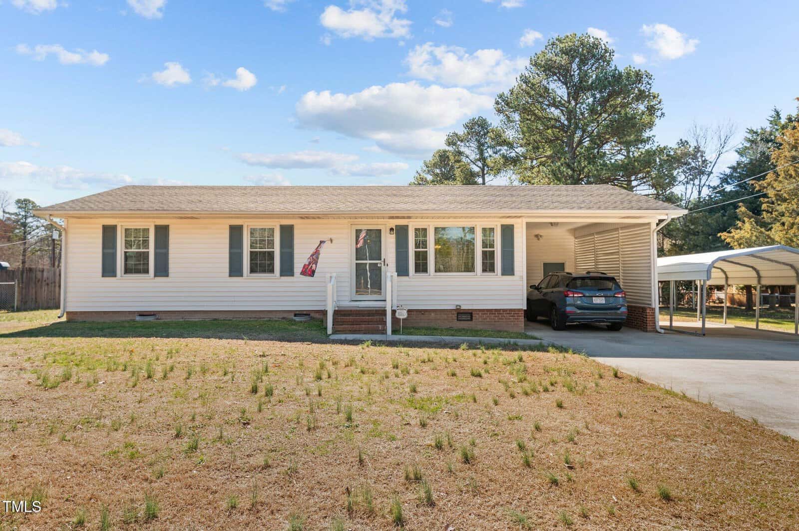 A single-story white house with brown shutters featuring a small front porch and an American flag. A driveway on the right leads to a carport with a parked car. The yard is sparse, with patches of grass. Tall trees are in the background under a blue sky.