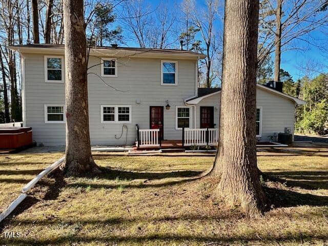 A two-story house with light-colored siding and several windows is visible. There is a small wooden deck with a railing in the back, surrounded by tall trees and a grassy yard. A hot tub is situated on the left side of the deck.