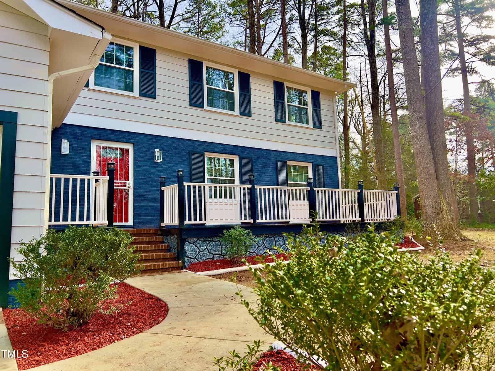 Situated at 1517 Peace Street in Henderson, this two-story house boasts a blue and white facade enveloped by trees. A striking red door and white railings greet you, while a walkway lined with shrubs and red mulch guides you to the welcoming steps.