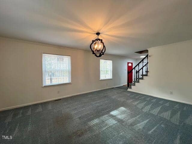 A spacious living room with light gray carpet and white walls. Two windows provide natural light. A black chandelier hangs from the ceiling. A staircase with black railing leads upstairs, and a red door is visible in the background.