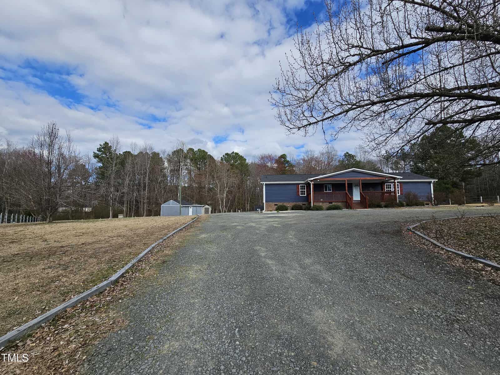 A gravel driveway leads to a single-story house with a dark exterior and a covered porch. The yard is expansive with grass and trees. A small shed is visible to the left. The sky is partly cloudy.