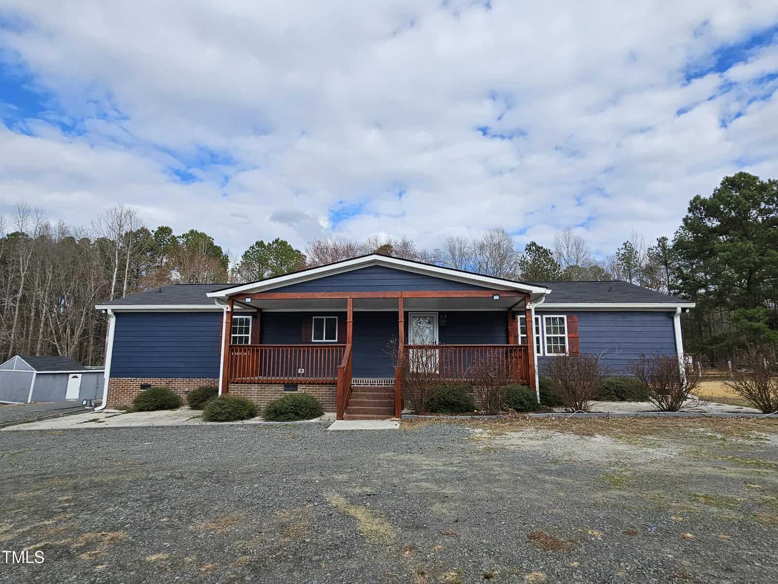 A single-story house with a blue exterior and a wooden front porch is set against a cloudy sky. A gravel driveway leads to the entrance. There is a small shed to the left and trees in the background.