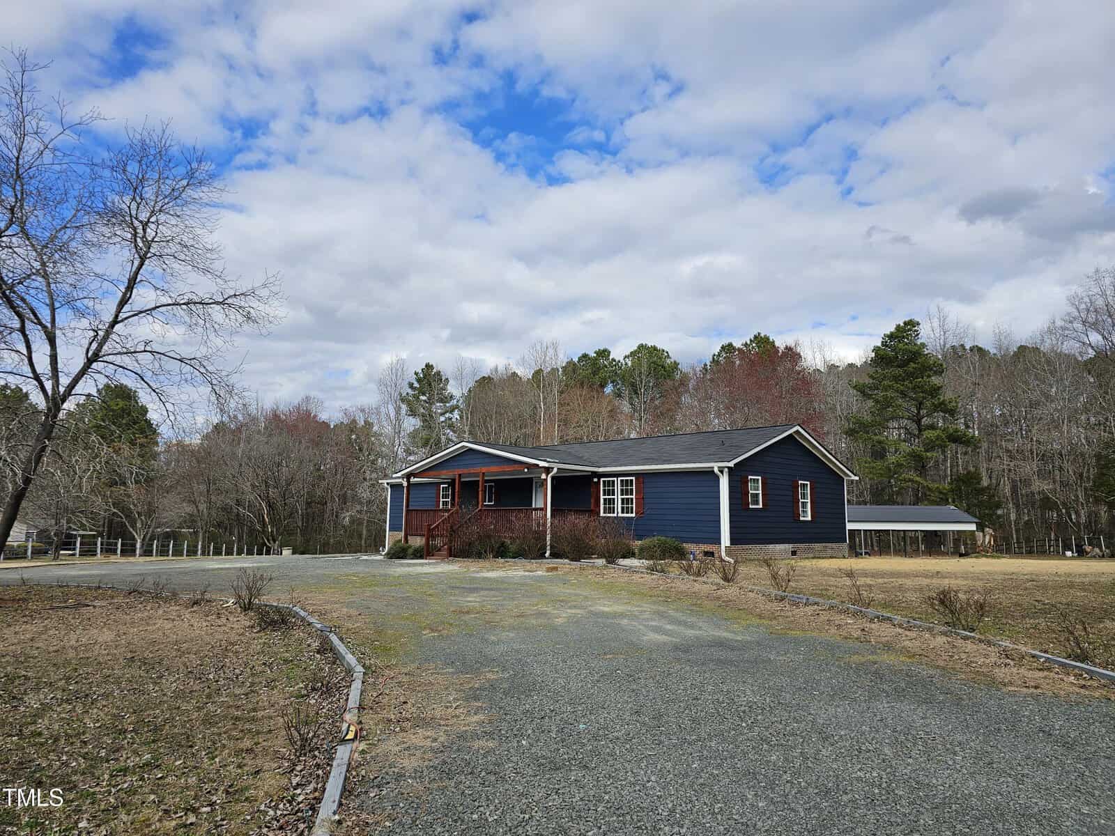 Nestled on 2126 Victory Lane, this charming single-story blue house in Creedmoor features a cozy covered porch by a gravel driveway. A lush lawn with bare trees encircles the home, while the partly cloudy sky sets a serene backdrop against a distant line of trees.