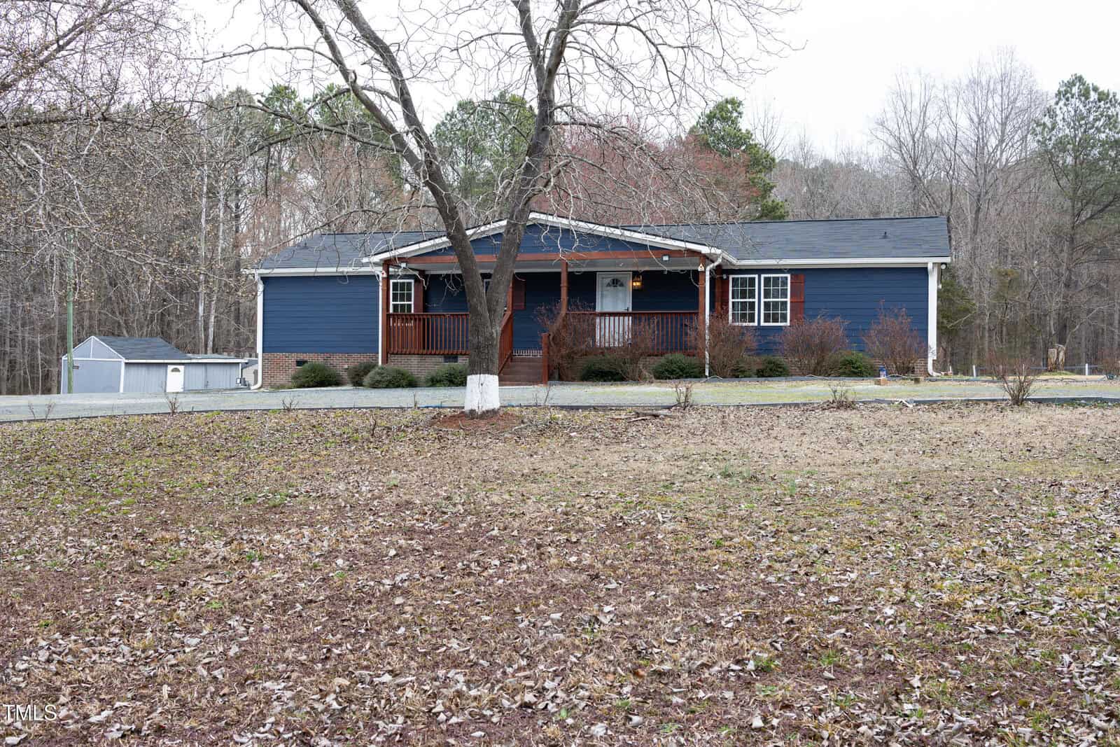A single-story blue house with a wooden porch is situated in a wooded area. The yard is covered with fallen leaves, and a bare tree stands in front. A small shed is visible to the left, and the background features trees with sparse foliage.