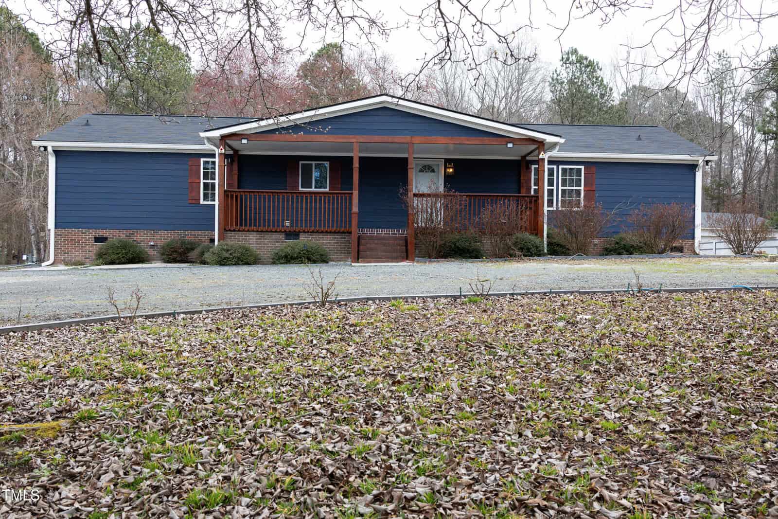 A blue ranch-style house with a brick foundation and white trim. It has a porch with wooden railings and a gravel driveway in front. The yard is bordered by trees, and the ground is covered with dry leaves.