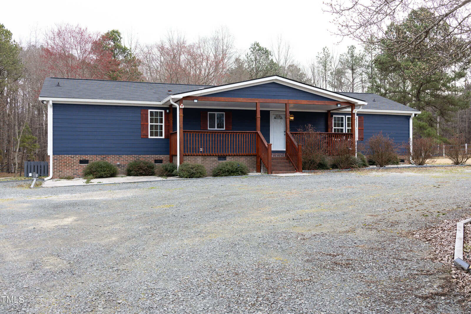 A blue, single-story house with a gabled roof and a covered front porch supported by wooden posts. It is surrounded by trees and shrubbery, with a gravel driveway in the foreground and a grassy area nearby. Overcast sky in the background.