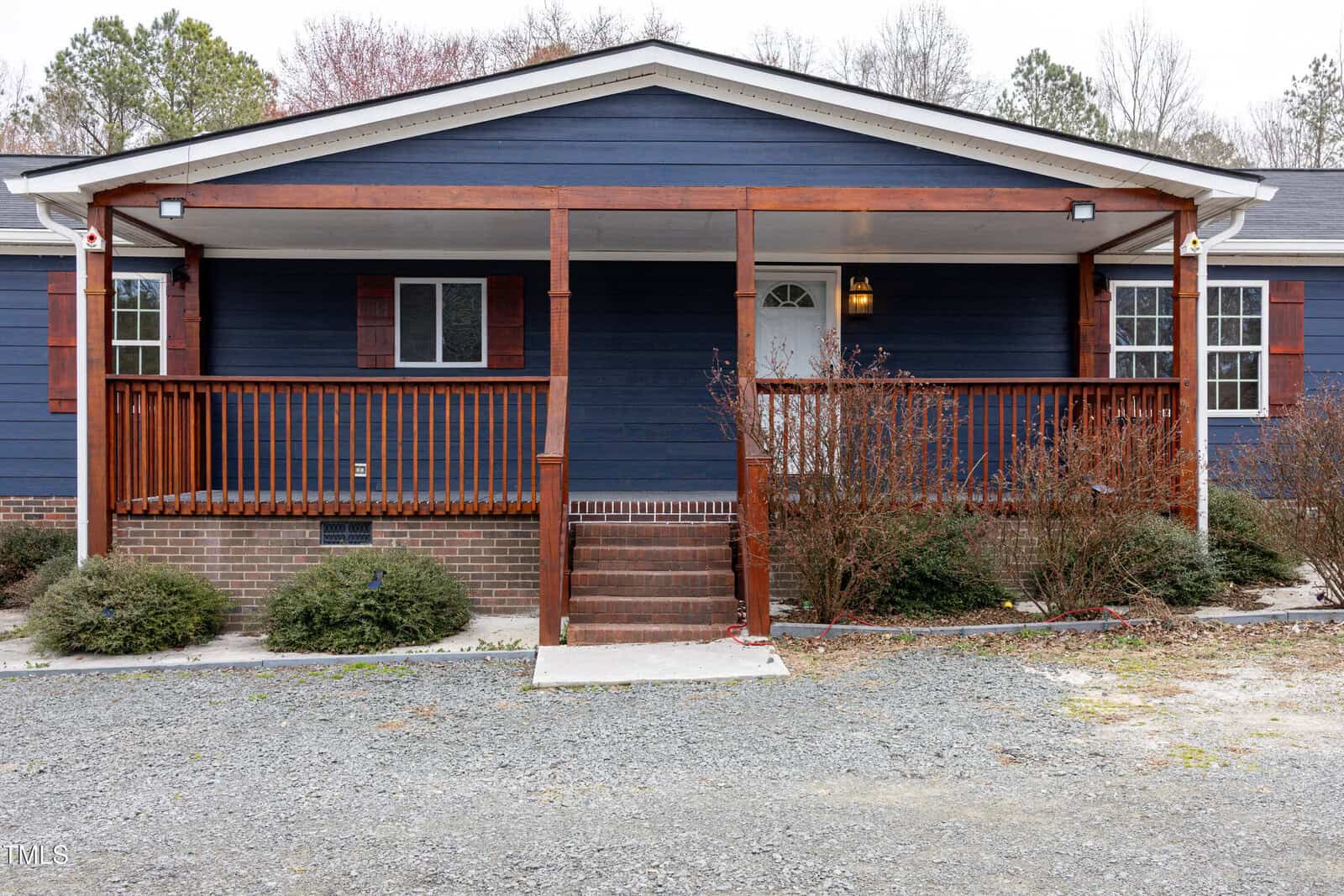 A blue single-story house with a brick foundation and wooden front porch. The porch has railings and two sets of steps. The front door is centered with a small window. Sparse bushes and a gravel driveway are in front. Trees are visible in the background.