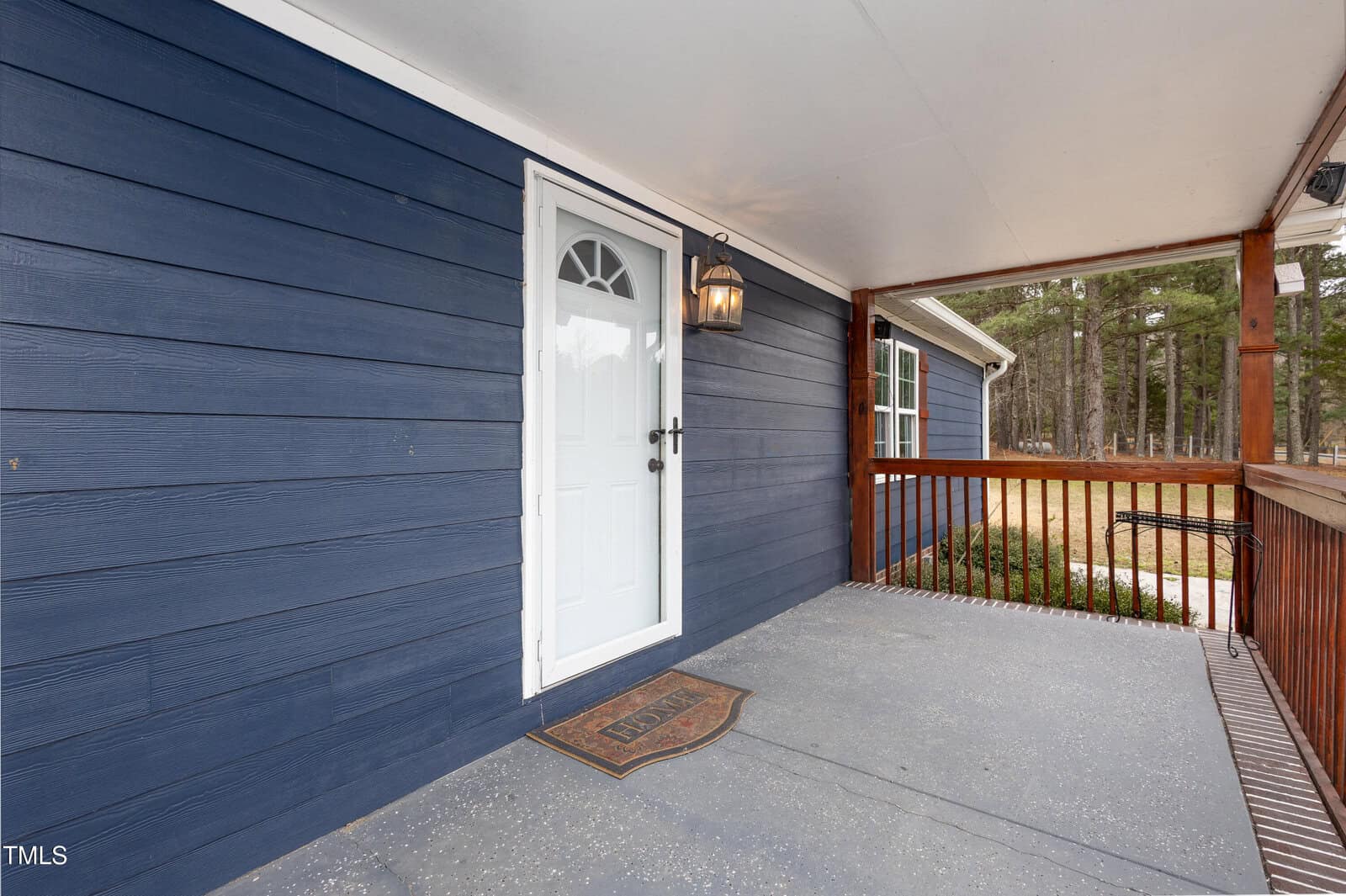 A blue house exterior features a white door with an arched window. A hanging lantern light is on the wall next to the door. The wooden porch has a mat labeled "HOME" and is enclosed by a wooden railing. Trees are visible in the background.