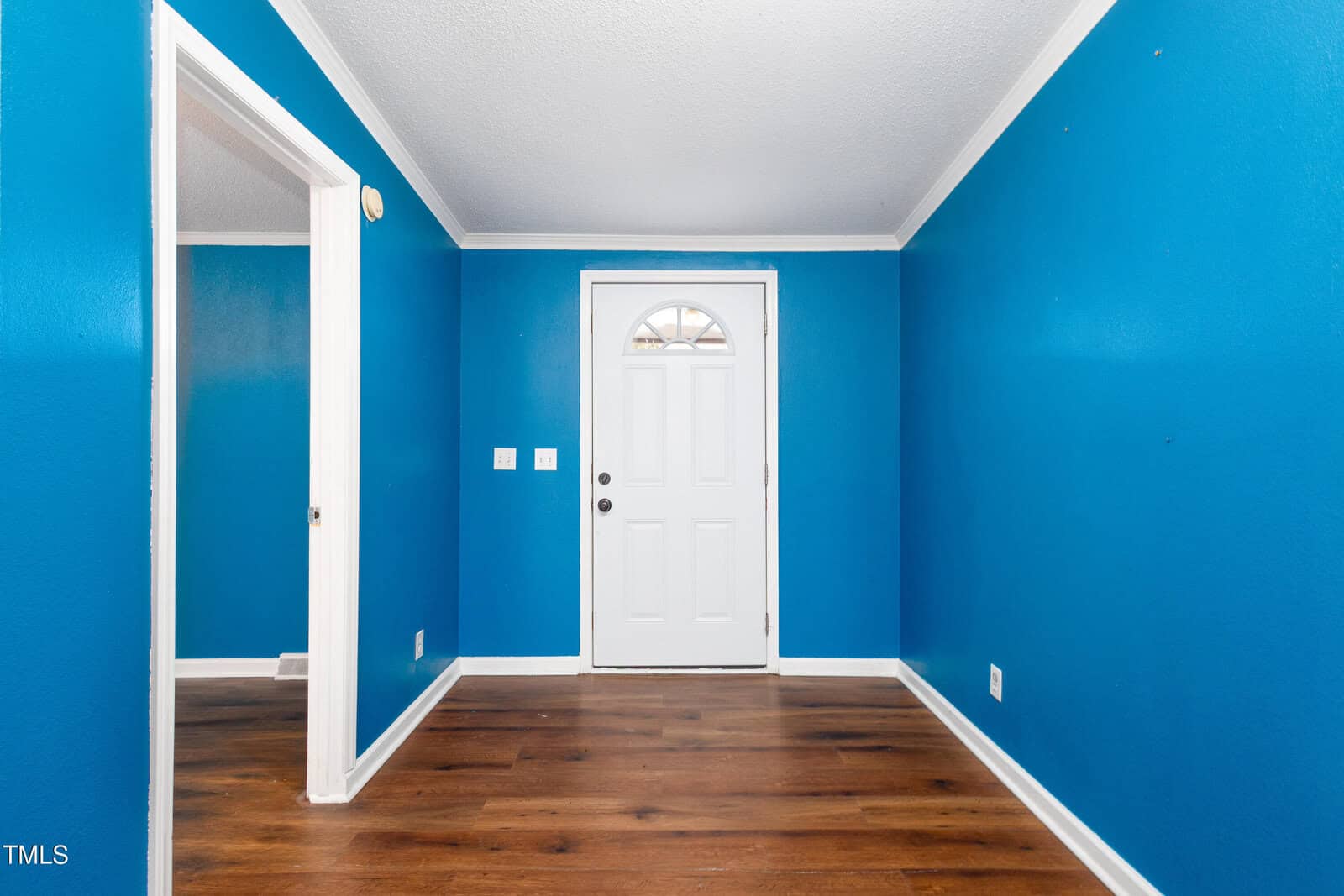 A small, empty room with bright blue walls and a wooden floor features a white front door with a semi-circular window at the top. To the left, there is an open doorway leading into another room with matching blue walls.