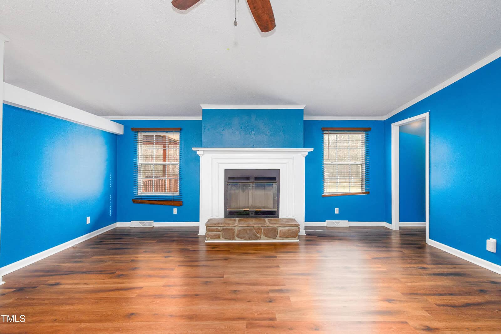 A living room with bright blue walls, a white fireplace centered on one wall, and two windows with wooden blinds. The floor is wood with a natural finish. A ceiling fan with wooden blades is visible above.